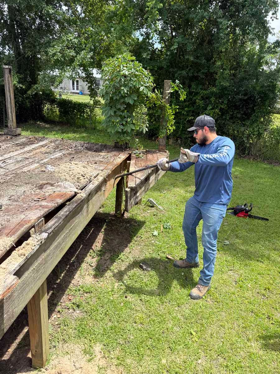 Worker using a pry bar to remove boards from a wooden deck outdoors.