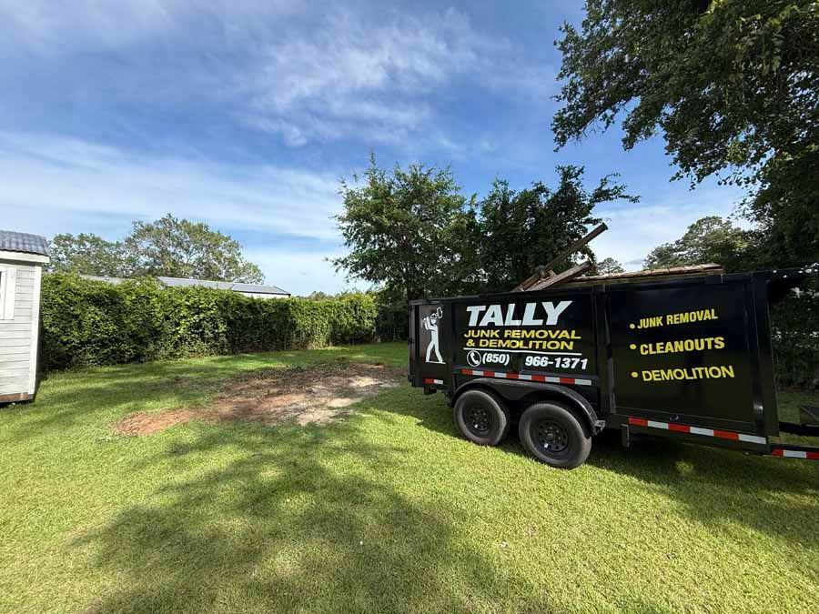 Black Tally landscaping trailer in a grassy yard beside trees and a white shed under a blue sky