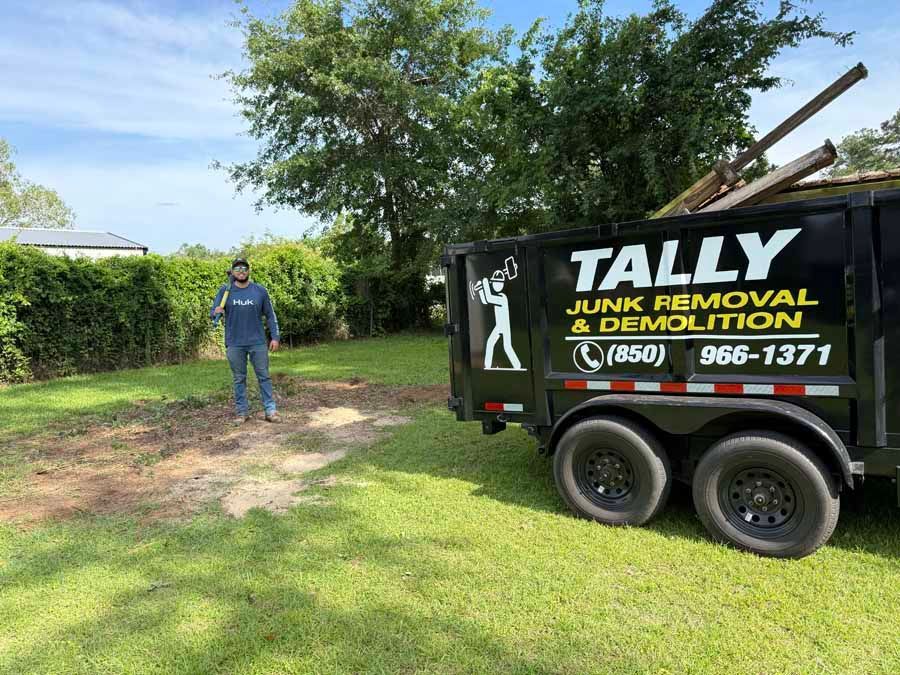 Man beside black Tally Junk Removal trailer in a sunny grassy yard with trees and hedges