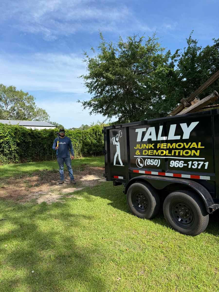 Man standing beside a Tally site demolition trailer in a grassy yard under a blue sky