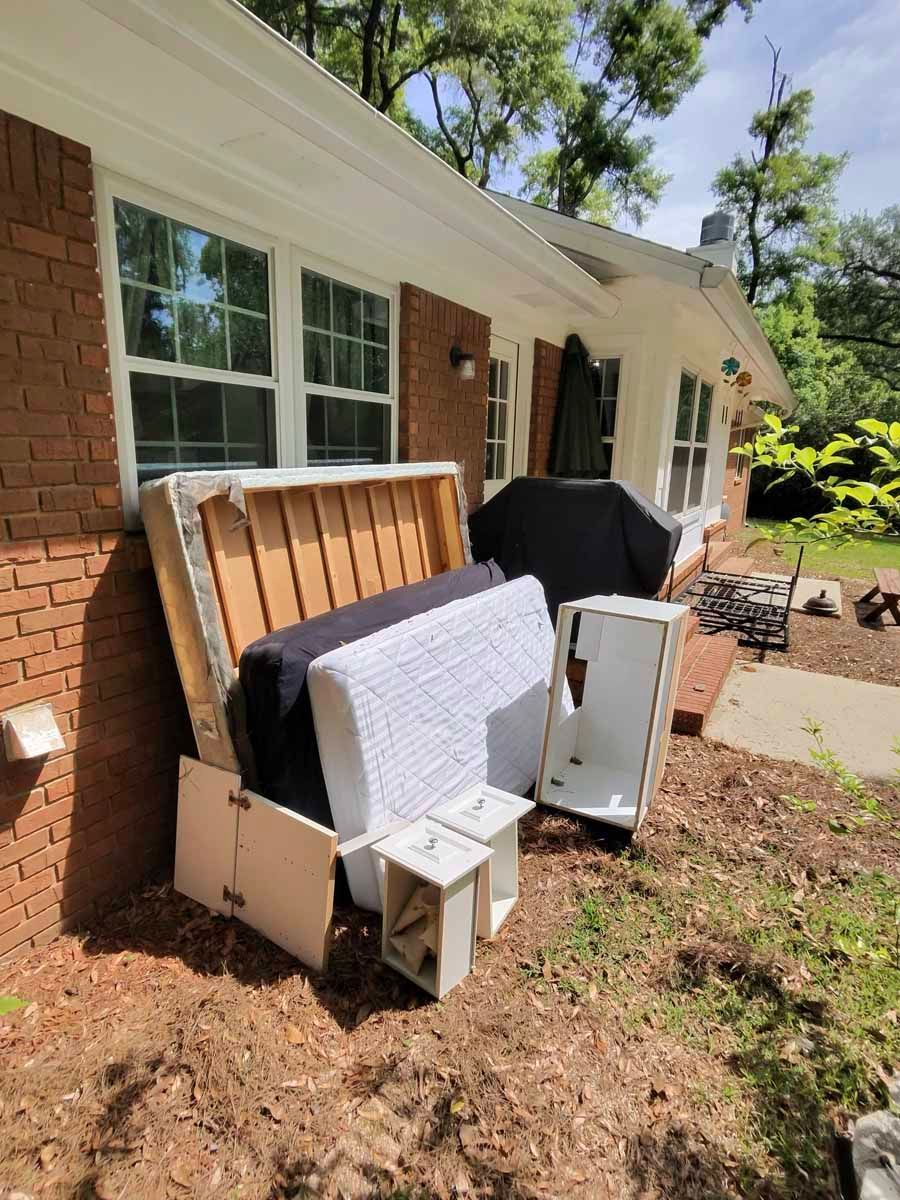 Discarded furniture and mattresses stacked beside a brick house in a yard.