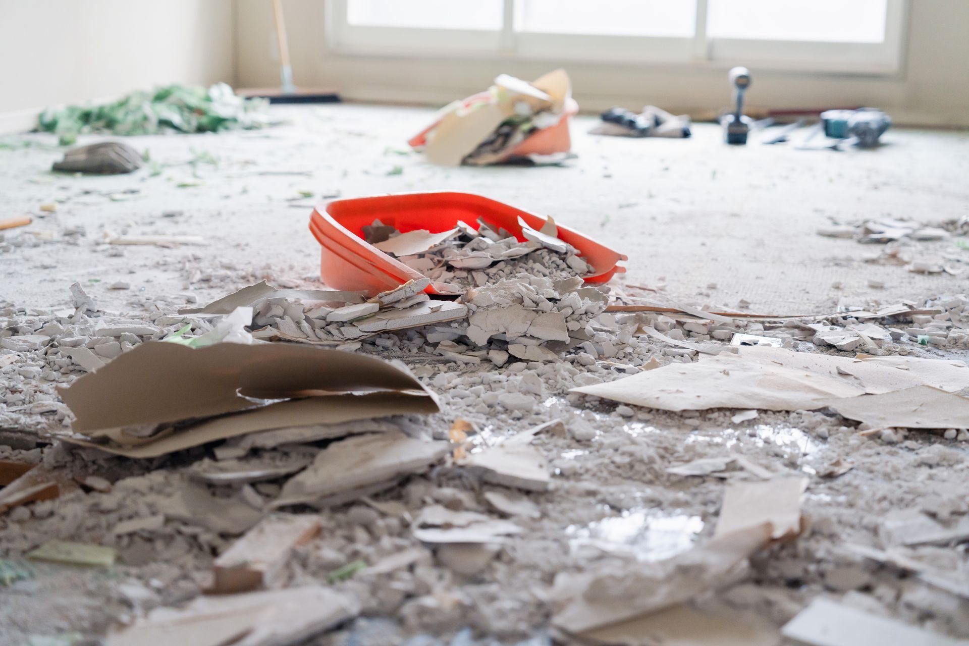 Debris and shattered drywall strewn across a room floor near a red bucket by a window