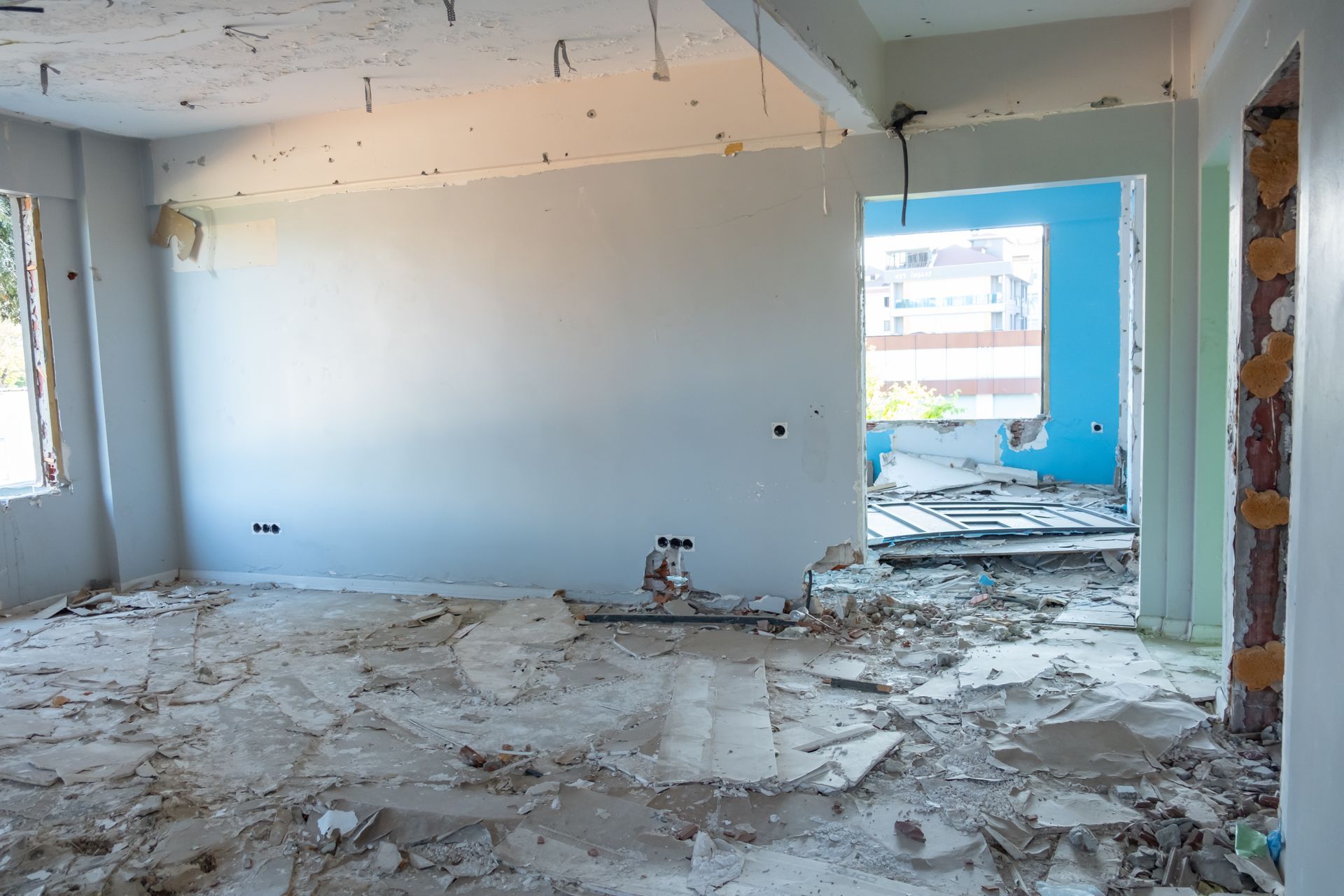 Damaged room with debris on the floor, broken walls, and a blue accent wall near a window.
