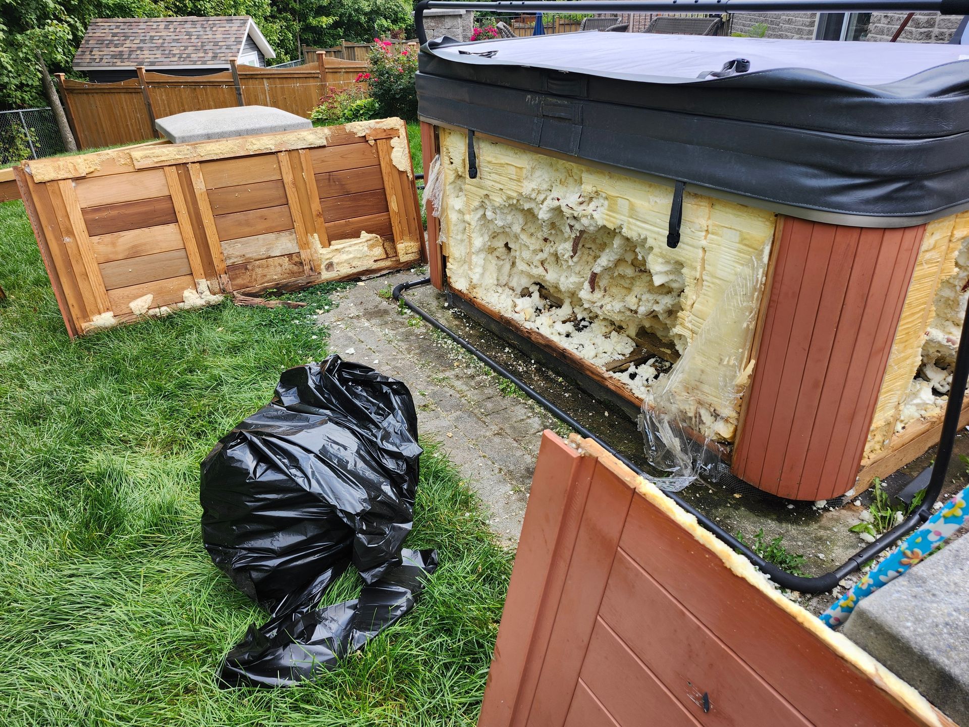 Damaged outdoor hot tub with peeled insulation, wooden panels, and black trash bags on grass