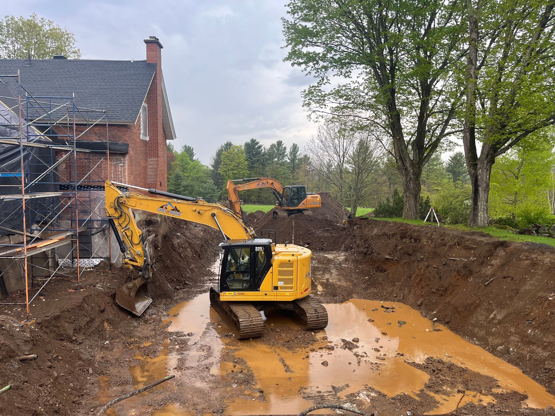 Une excavatrice jaune creuse un trou dans la terre devant une maison.