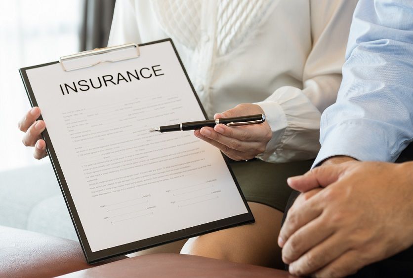 Woman points to an insurance document on a clipboard, reviewing with a seated man.
