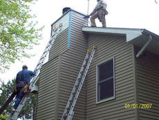 Two workers on ladders repairing a chimney on a two-story house with brown siding.