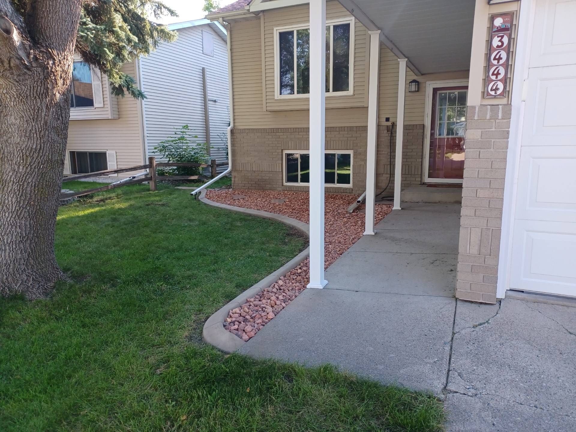Tan house front exterior with a concrete walkway, red landscaping rocks, and a green lawn under a clear blue sky.