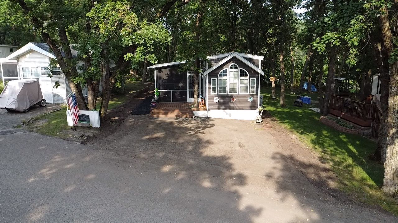 A small, gray cabin with a screened porch sits in a wooded area, accessed by a dirt path from a paved road.