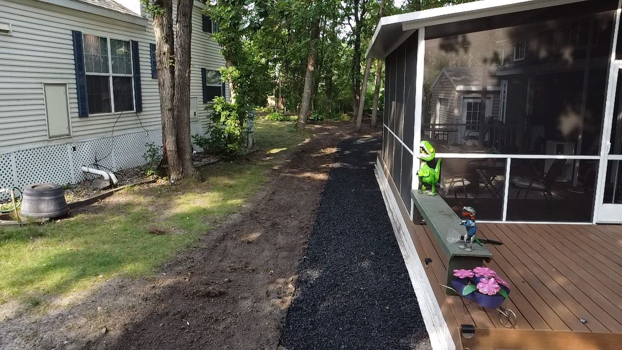 A strip of black rubber mulch sits along the side of a wooden deck next to an area of bare dirt and a white house.