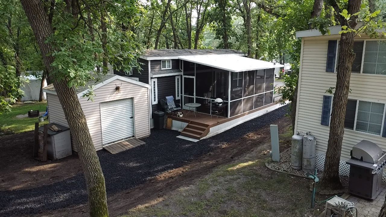 An elevated view of a small house with an attached screen porch and a separate garage, surrounded by trees and a dirt path.
