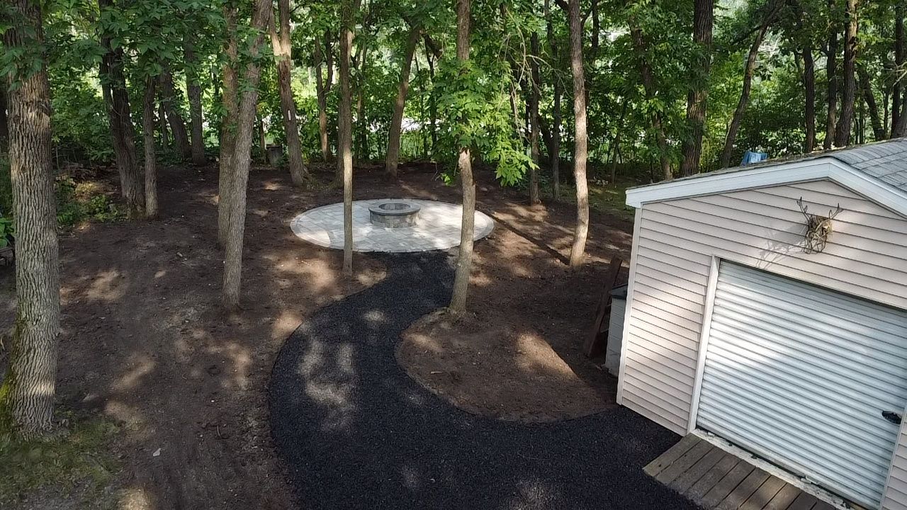 A dark gravel path leads from a shed to a circular gravel fire pit area in a wooded backyard.