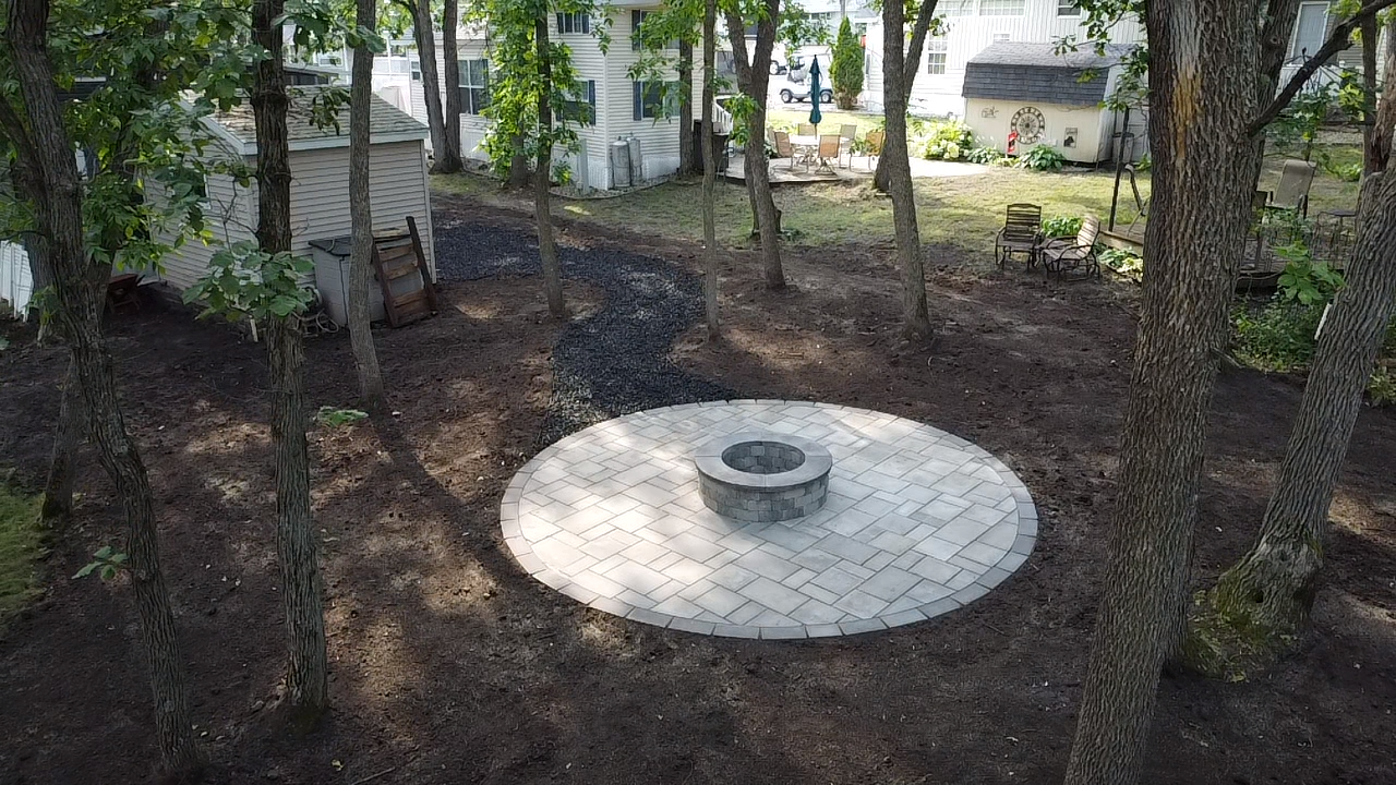 A circular light gray paver patio with a central stone fire pit, nestled in a wooded backyard near a garden shed.