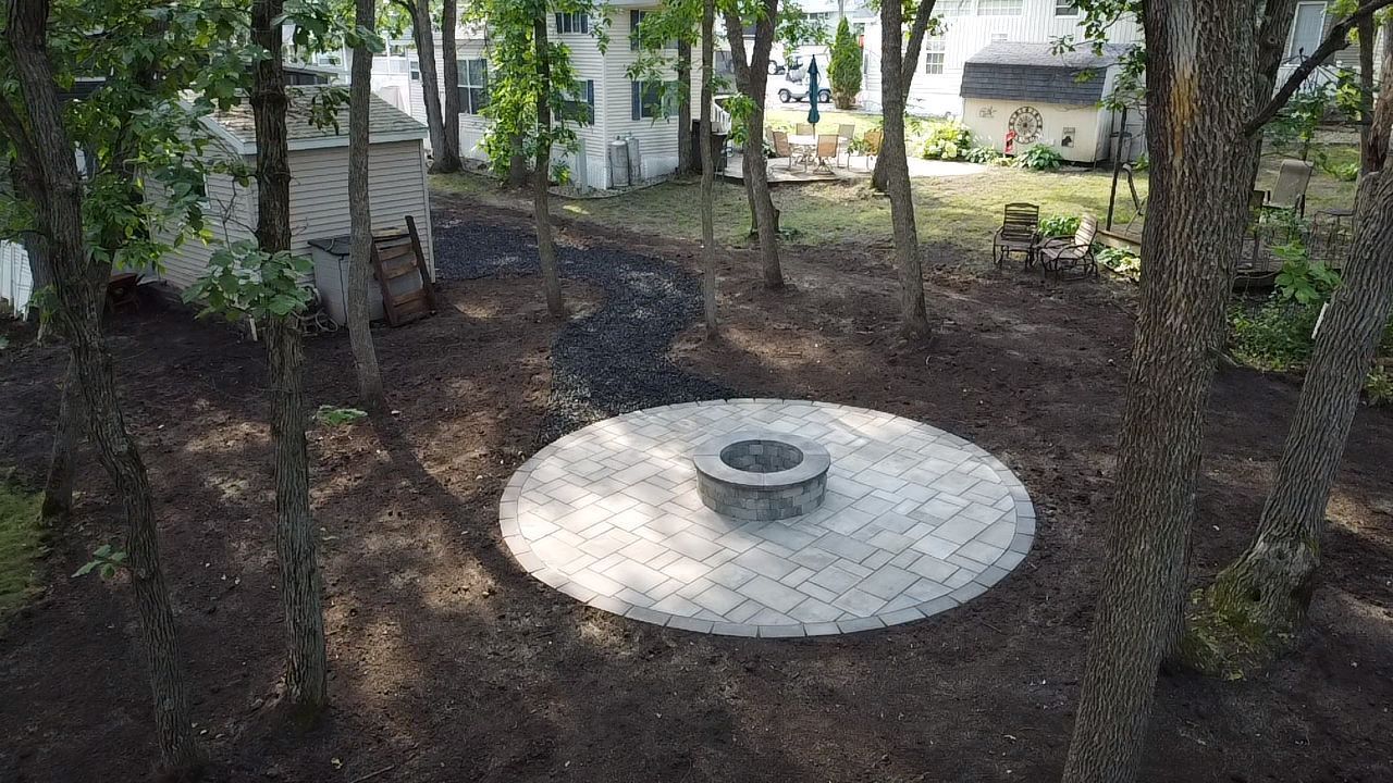 A circular stone paver patio with a fire pit in the center, surrounded by trees and dark mulch in a backyard.