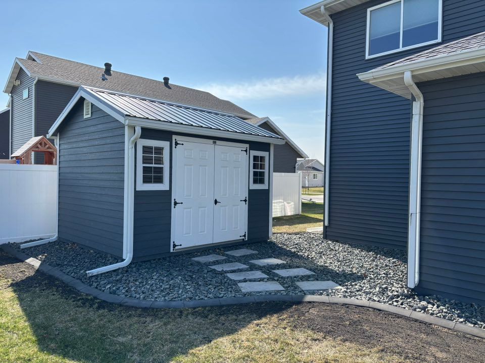 A dark blue storage shed with white trim and double doors sits on a gravel bed next to a house with matching siding.