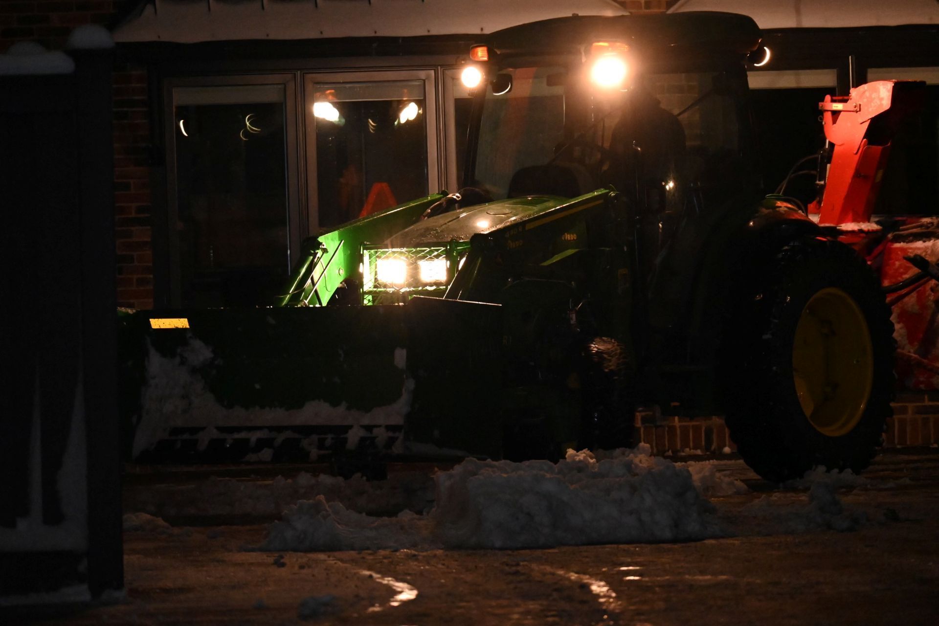 A green John Deere tractor with a plow clears snow in front of a building at night, illuminated by bright headlights.