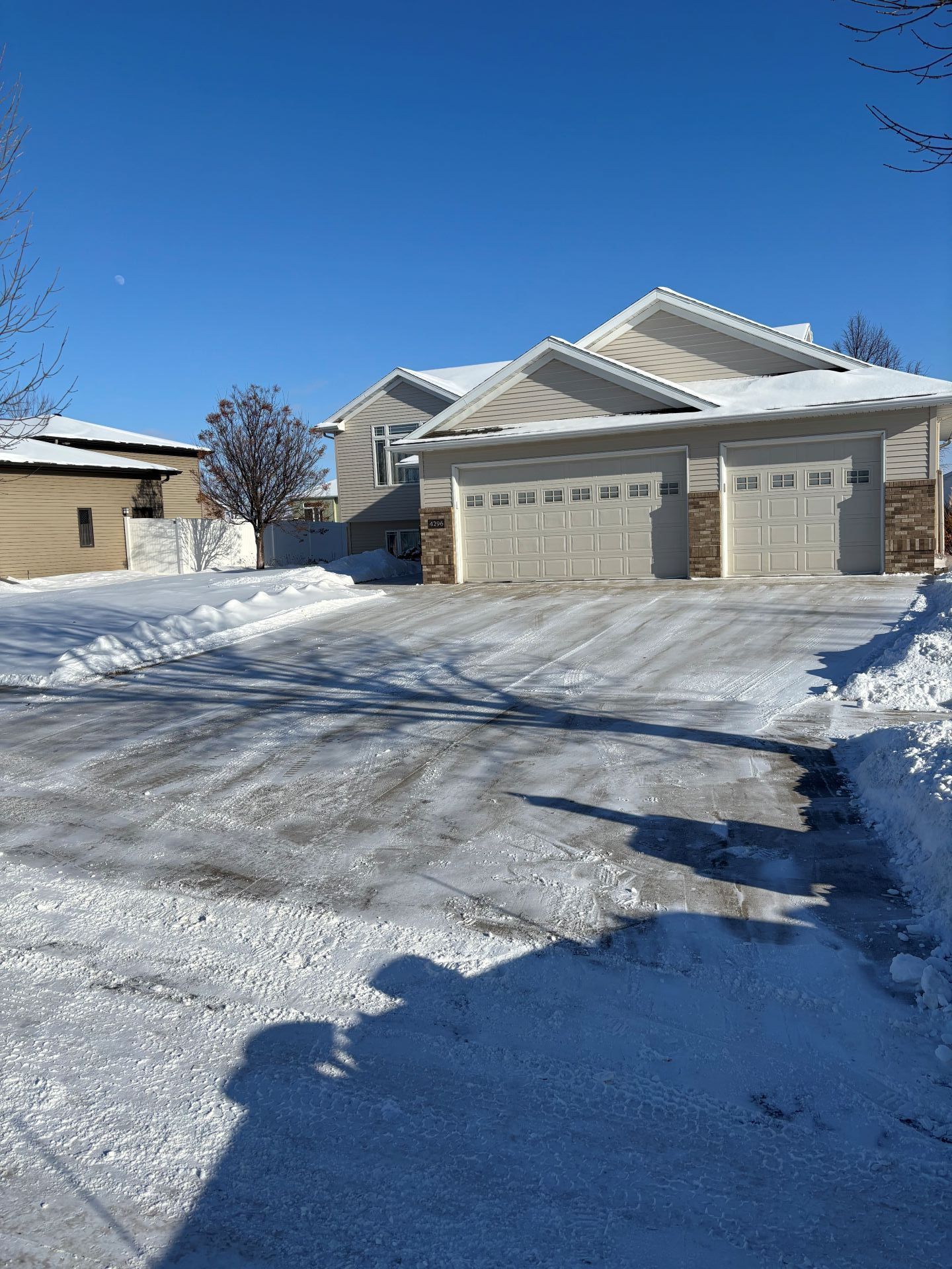 A tan house with a three-car garage, seen from a snow-covered driveway under a clear blue sky on a sunny winter day.