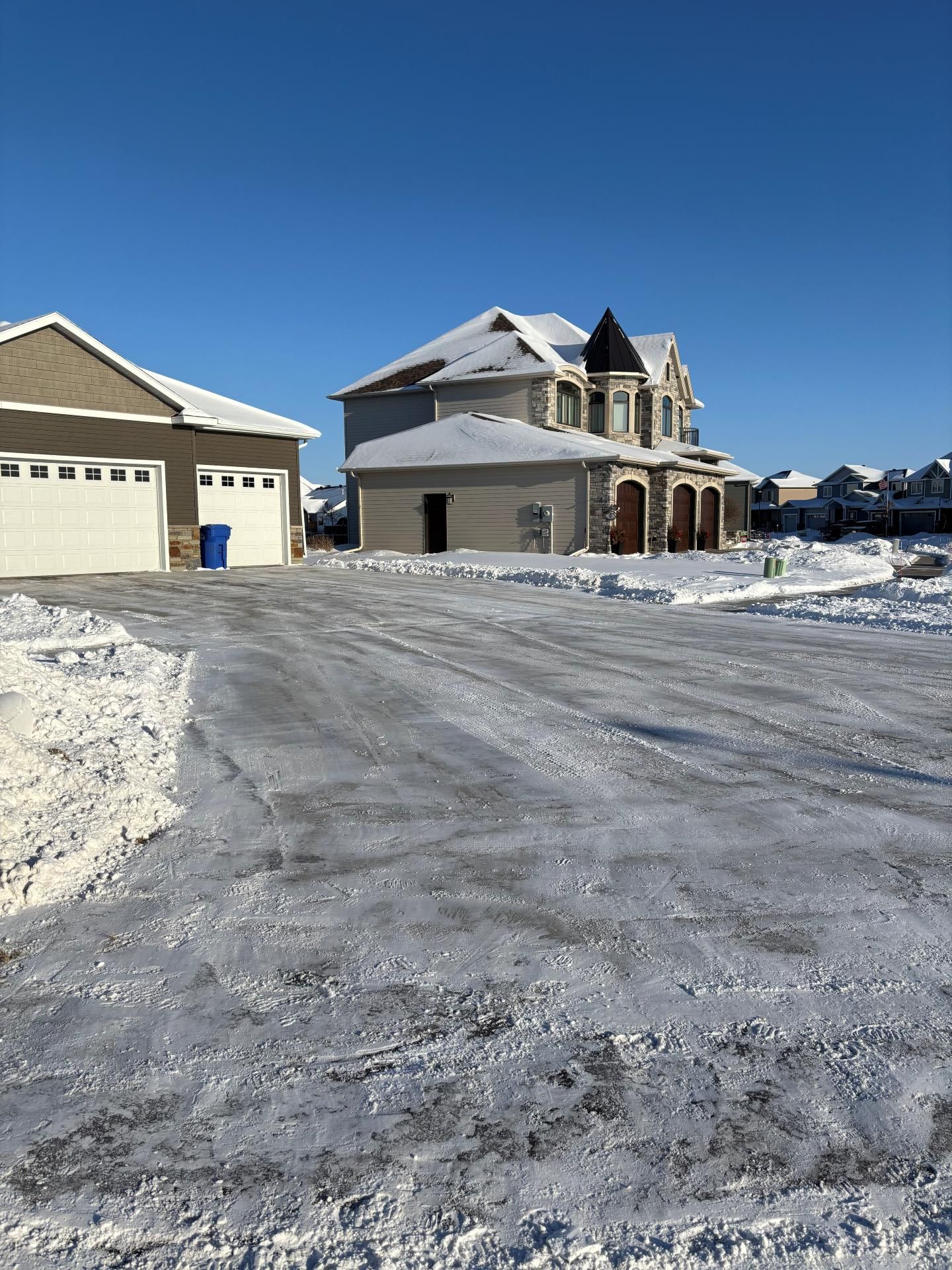 A beige house and garage under a clear blue sky, surrounded by snow-covered driveways and lawns.