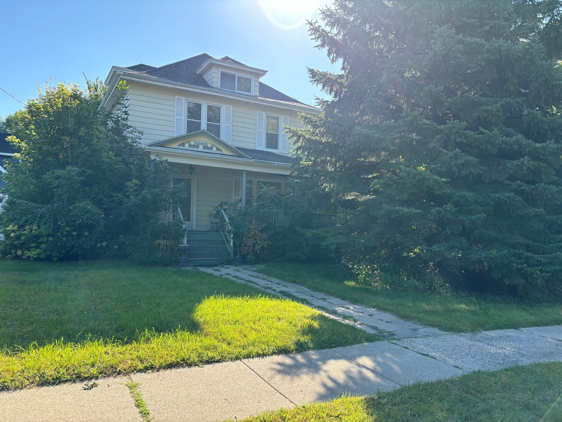 A two-story, cream-colored house with a front porch, framed by a large evergreen tree and smaller shrubs on a sunny day.
