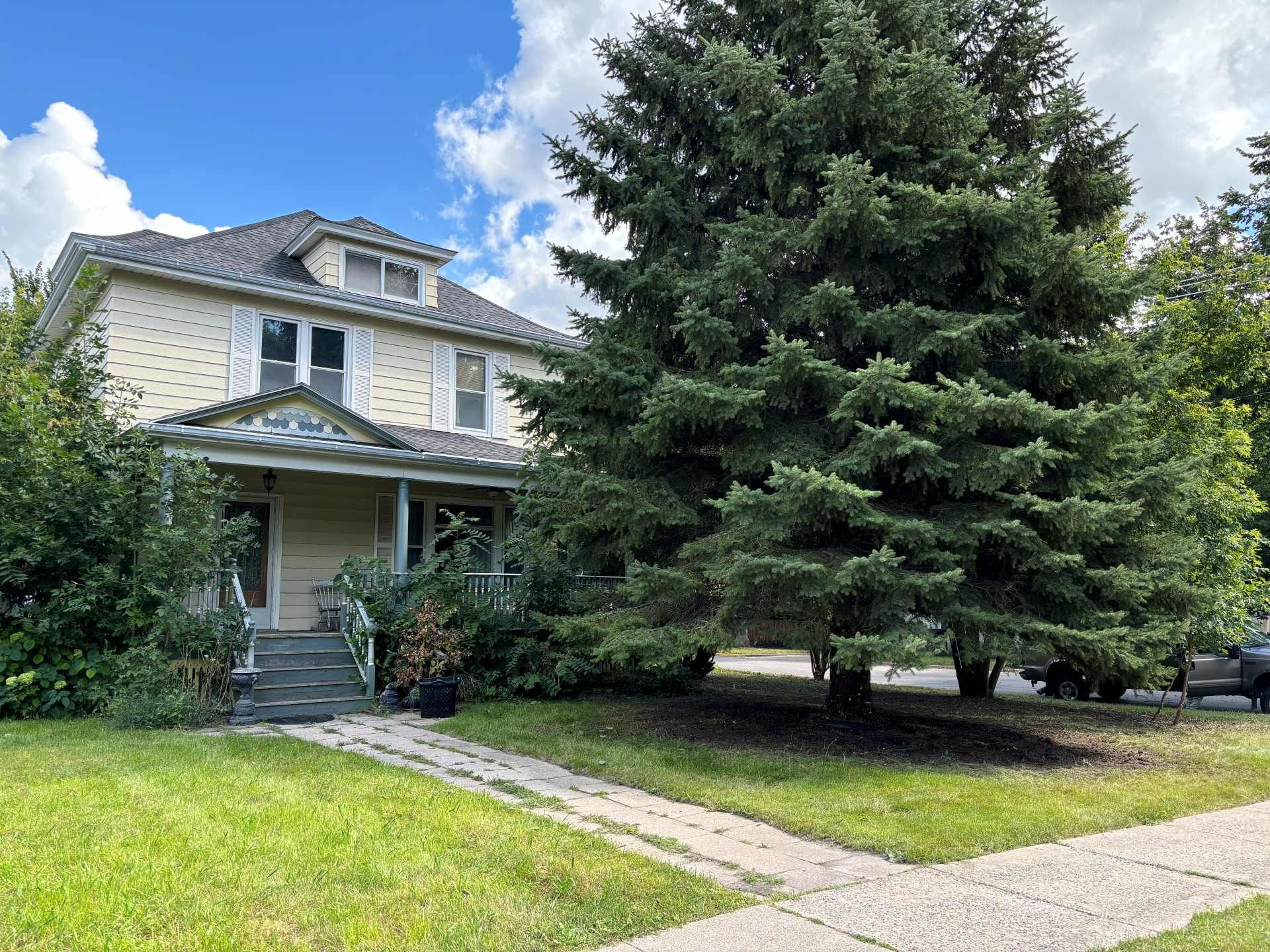 A two-story light-colored house with a front porch, surrounded by lush trees under a cloudy blue sky.