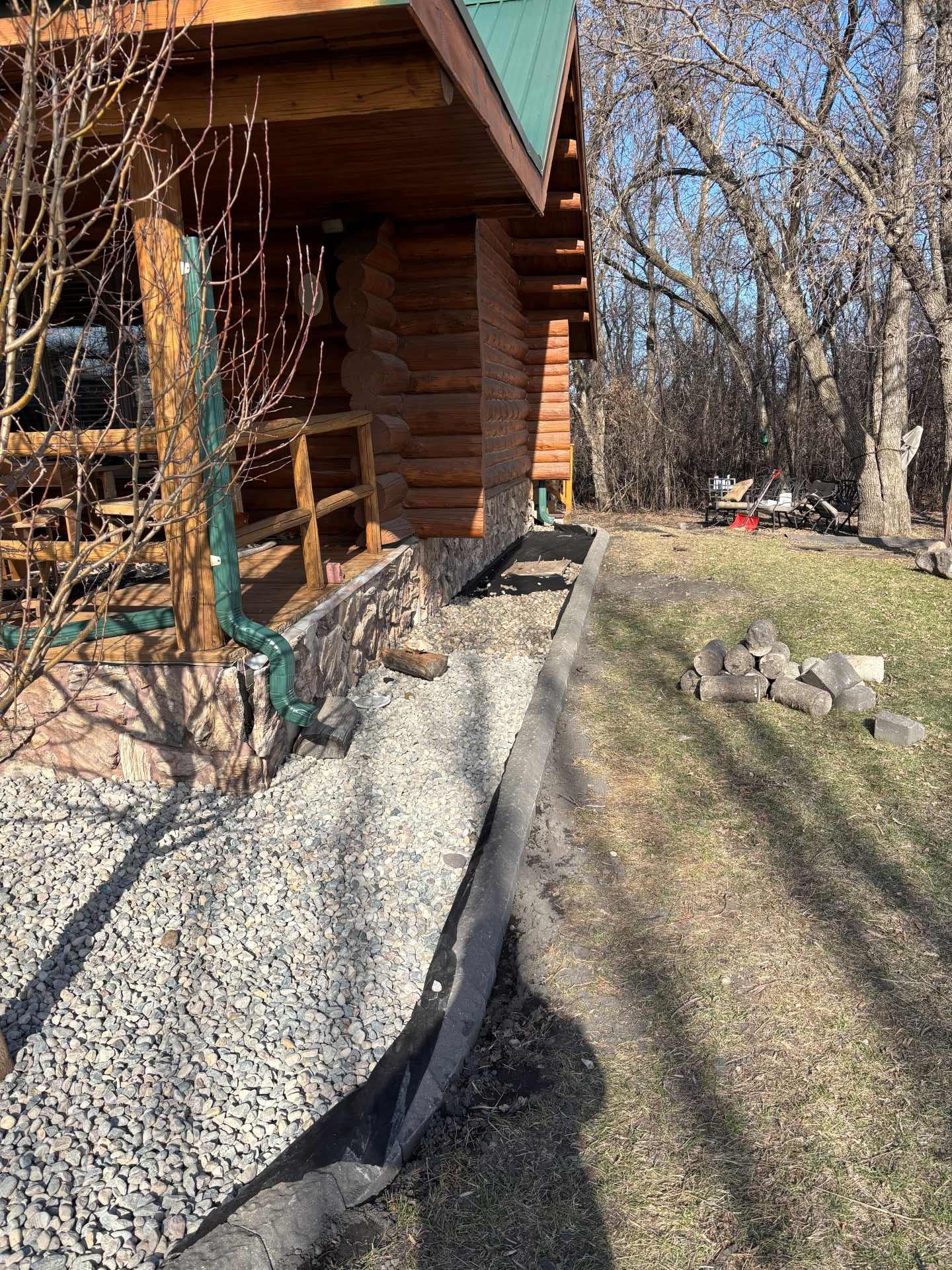 A log cabin porch with a gravel drainage border and black plastic edging, next to a grassy yard with trees.