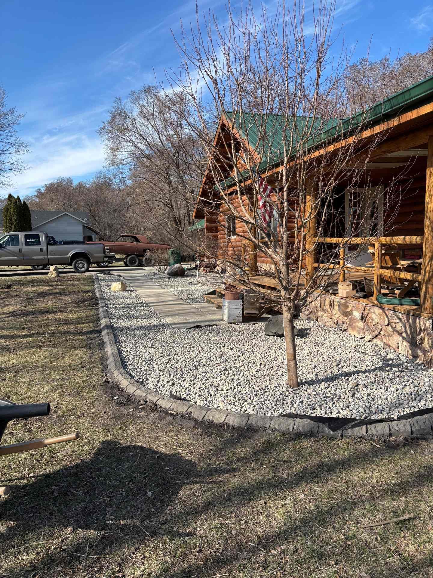 A log cabin with a stone-bordered gravel yard and a small tree in front, under a clear blue sky.
