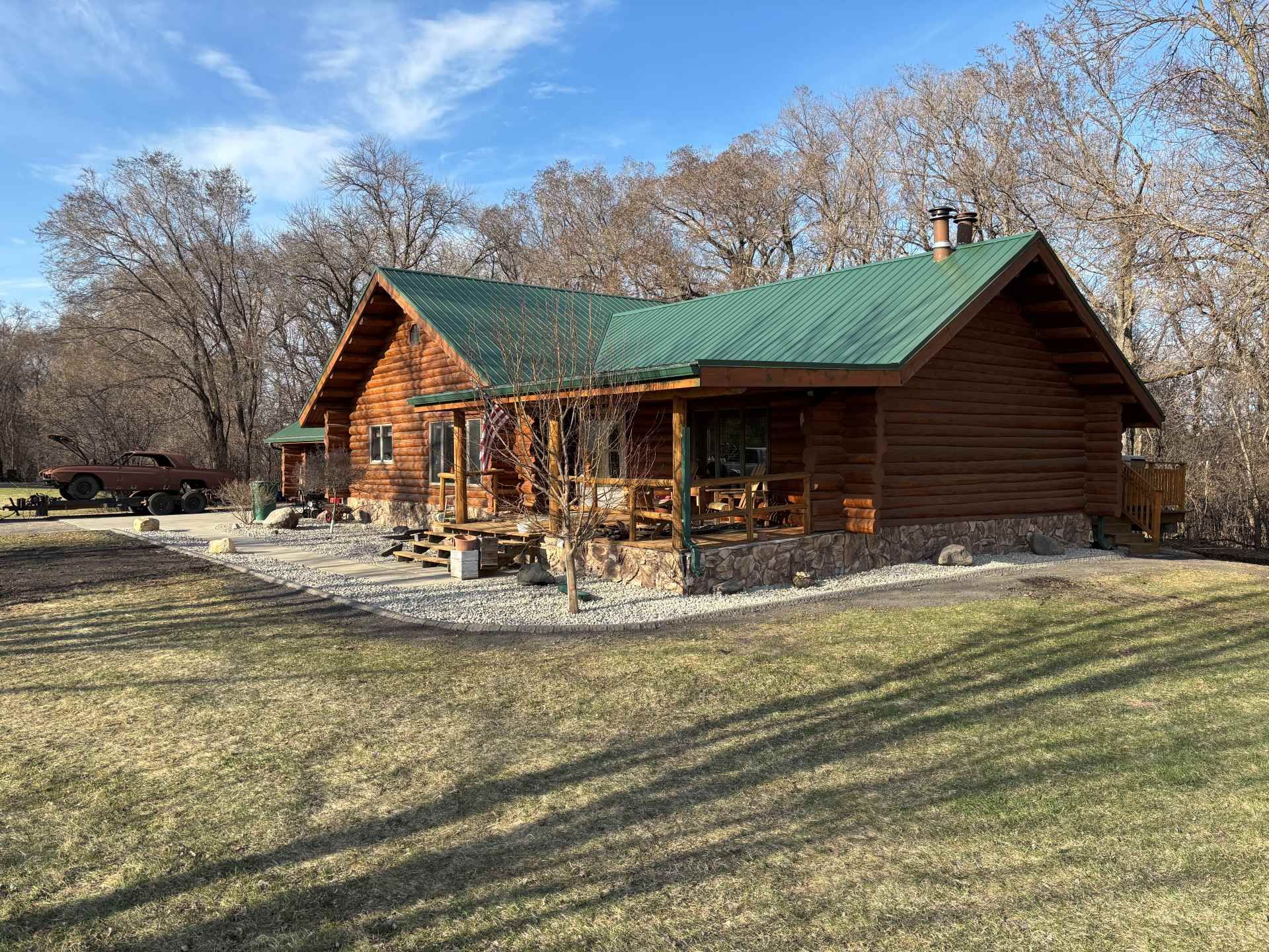 A rustic log cabin with a green metal roof, stone foundation, and covered porch, situated in a wooded, grassy yard.
