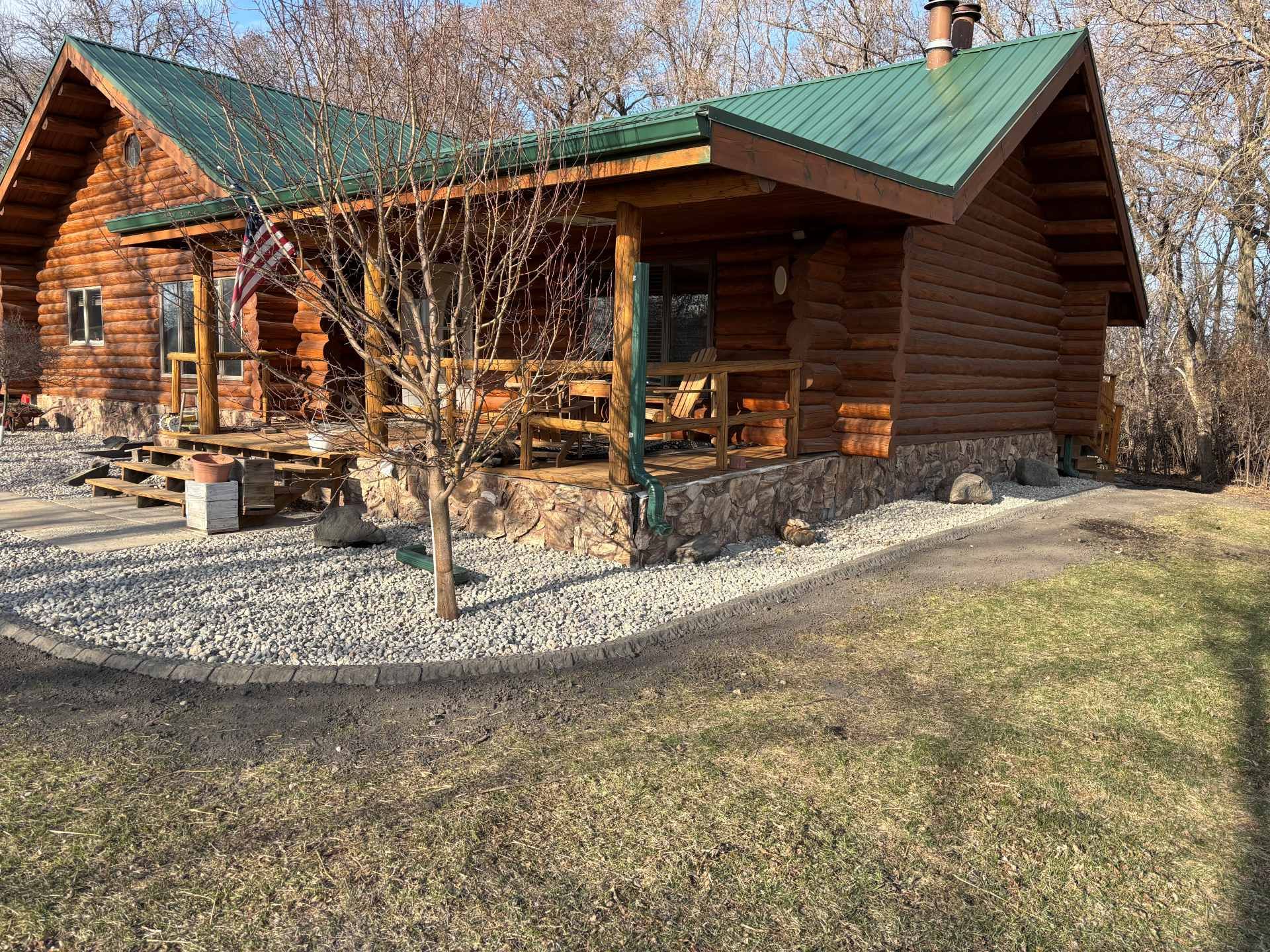 A rustic log cabin with a green metal roof and a stone foundation, surrounded by a gravel landscaping border and trees.