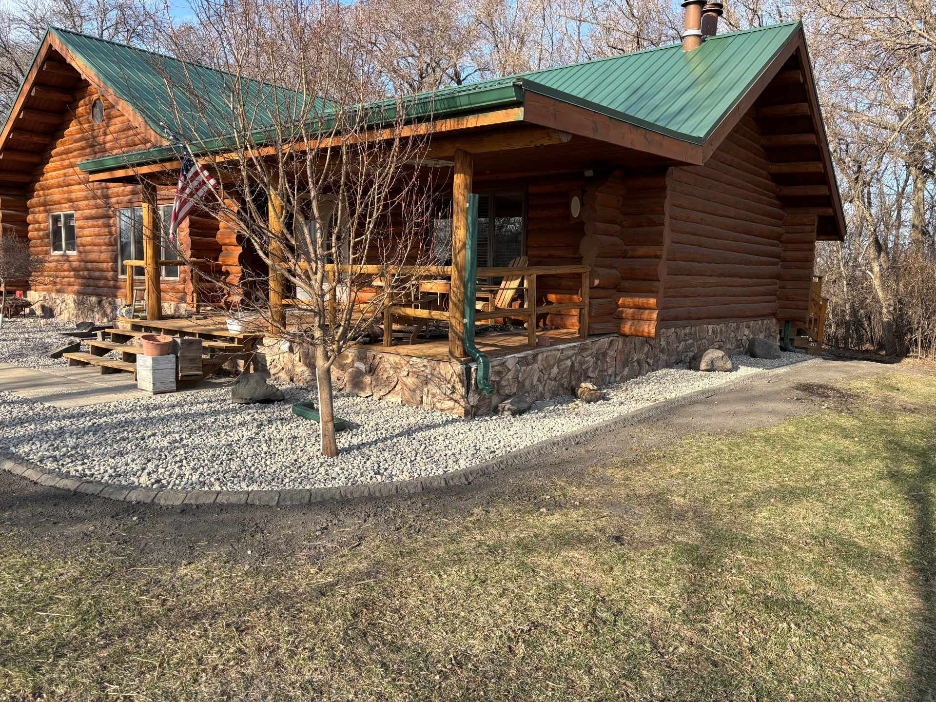 A rustic log cabin with a green metal roof, a wrap-around porch, and a stone foundation set among bare trees in winter.