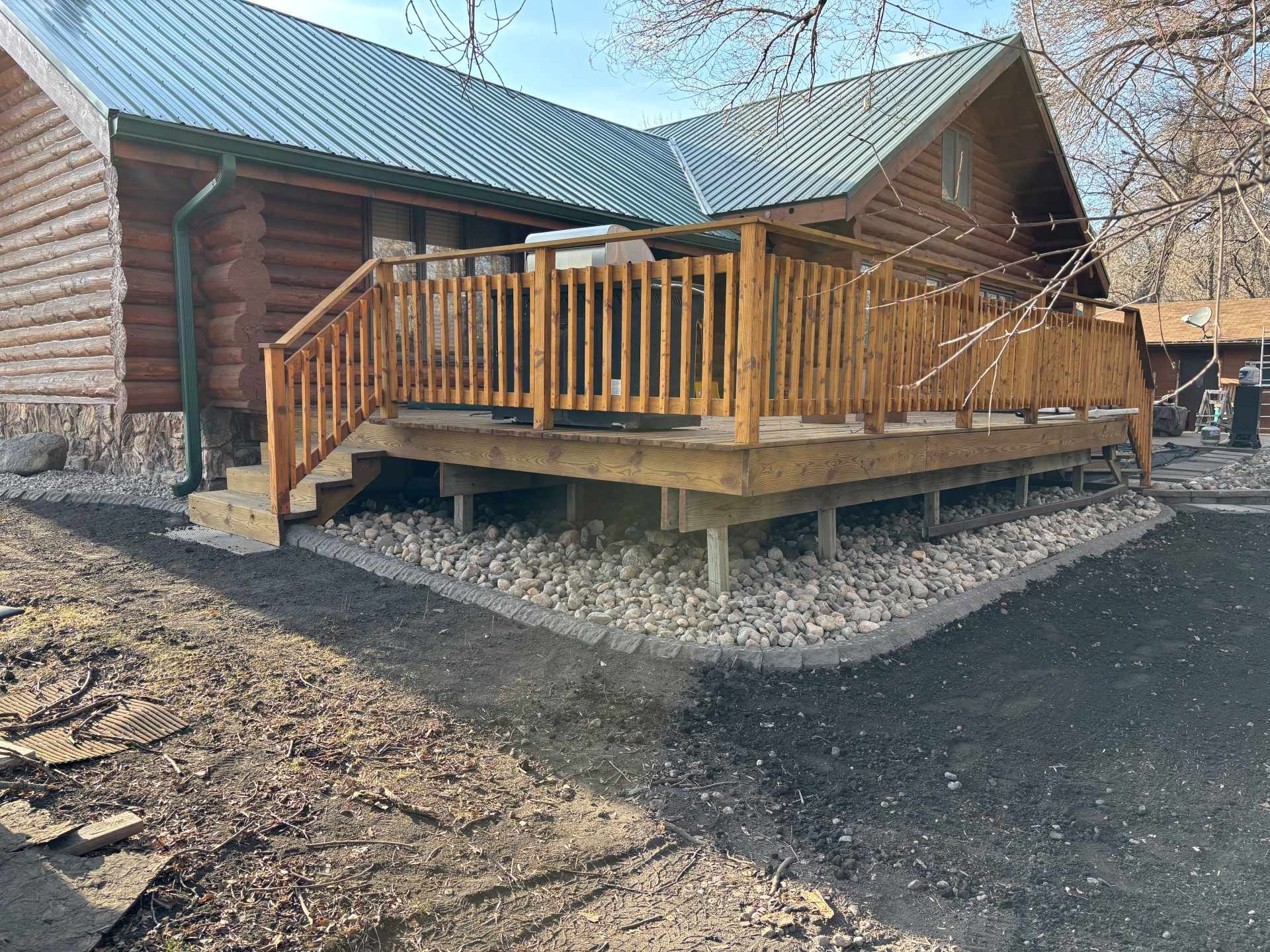 A rustic log cabin featuring a wooden deck with railing, surrounded by a light stone border and a dirt ground.