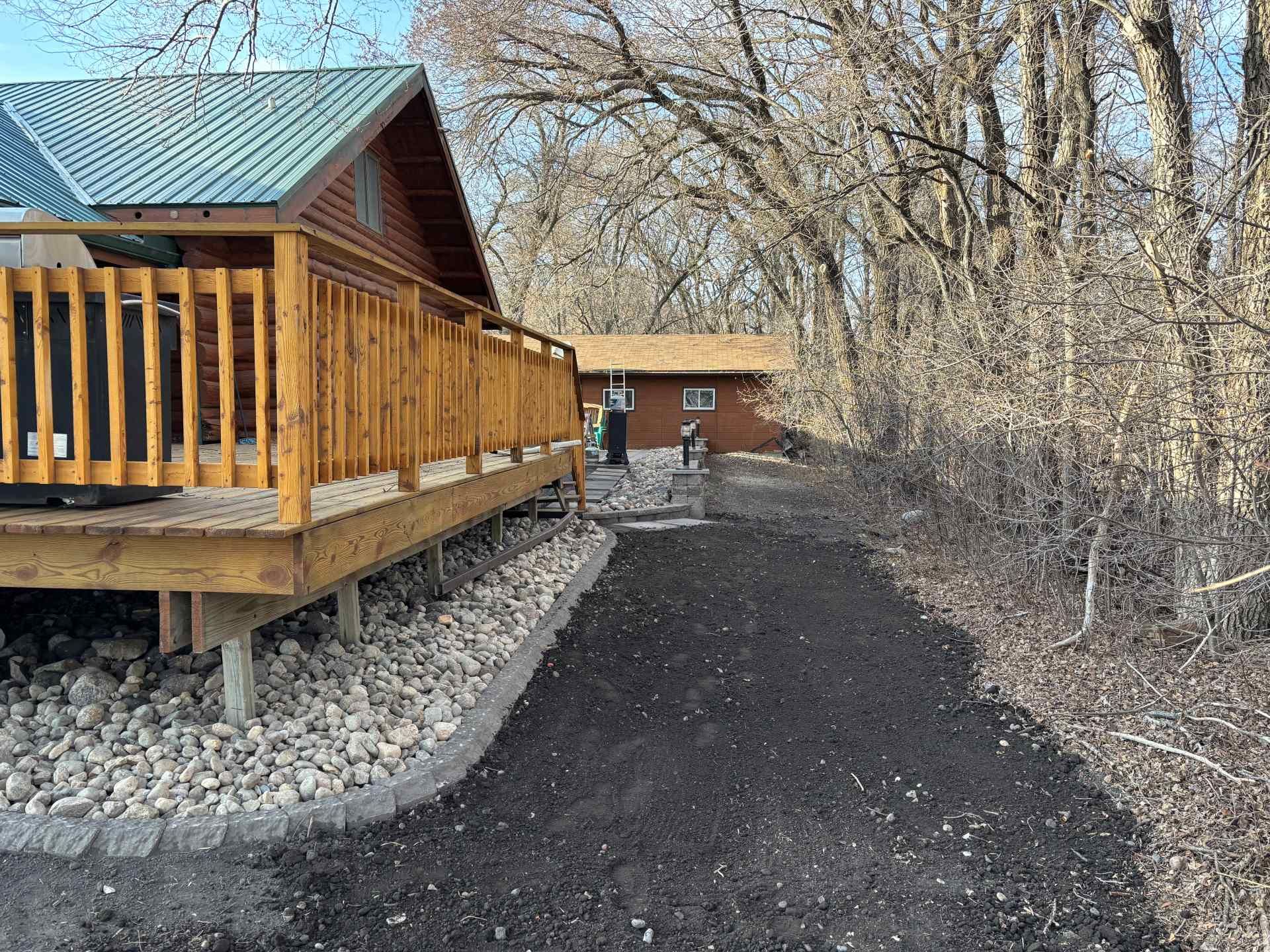 A wooden deck with a green roof sits beside a dark dirt path leading to a smaller cabin among bare, deciduous trees.