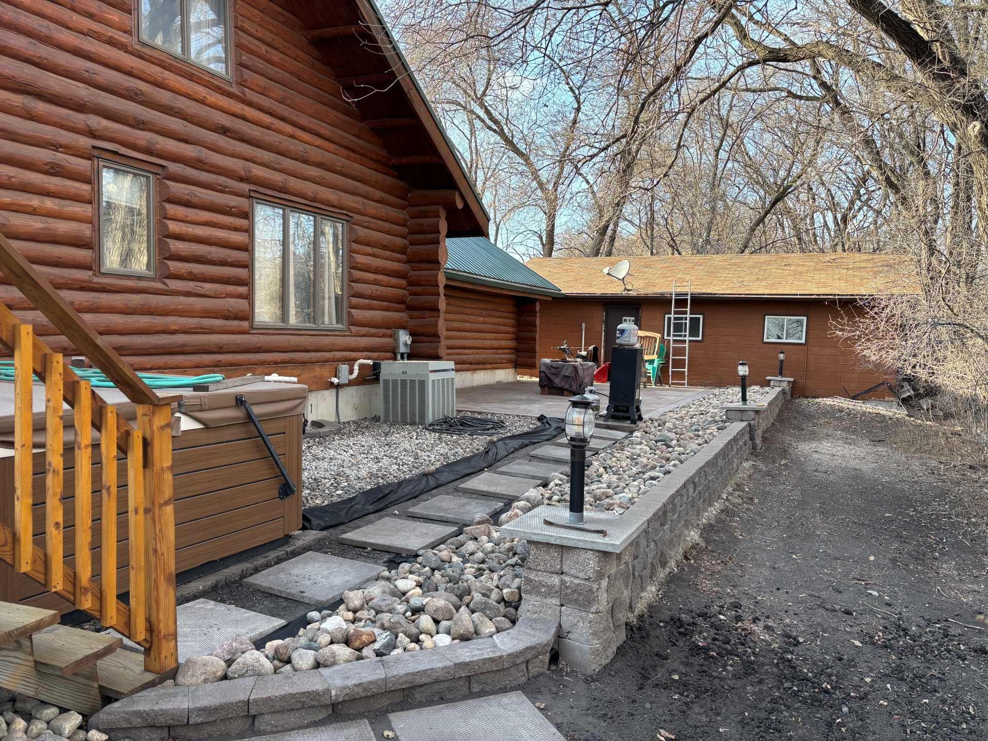 A brown log cabin exterior with a stone-paved walkway, rock garden, hot tub, and a detached building in the background.