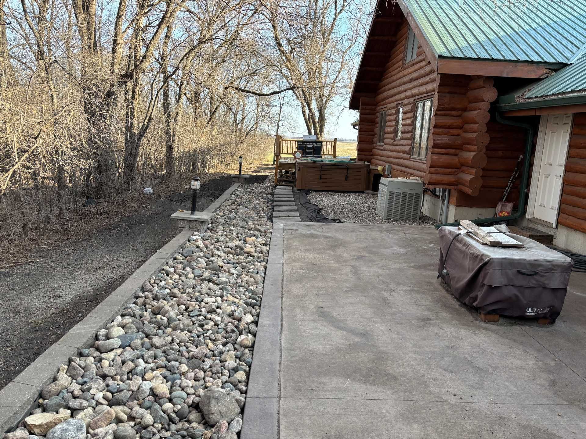 A concrete patio next to a log cabin, bordered by a long strip of decorative river rocks and a yard with bare trees.
