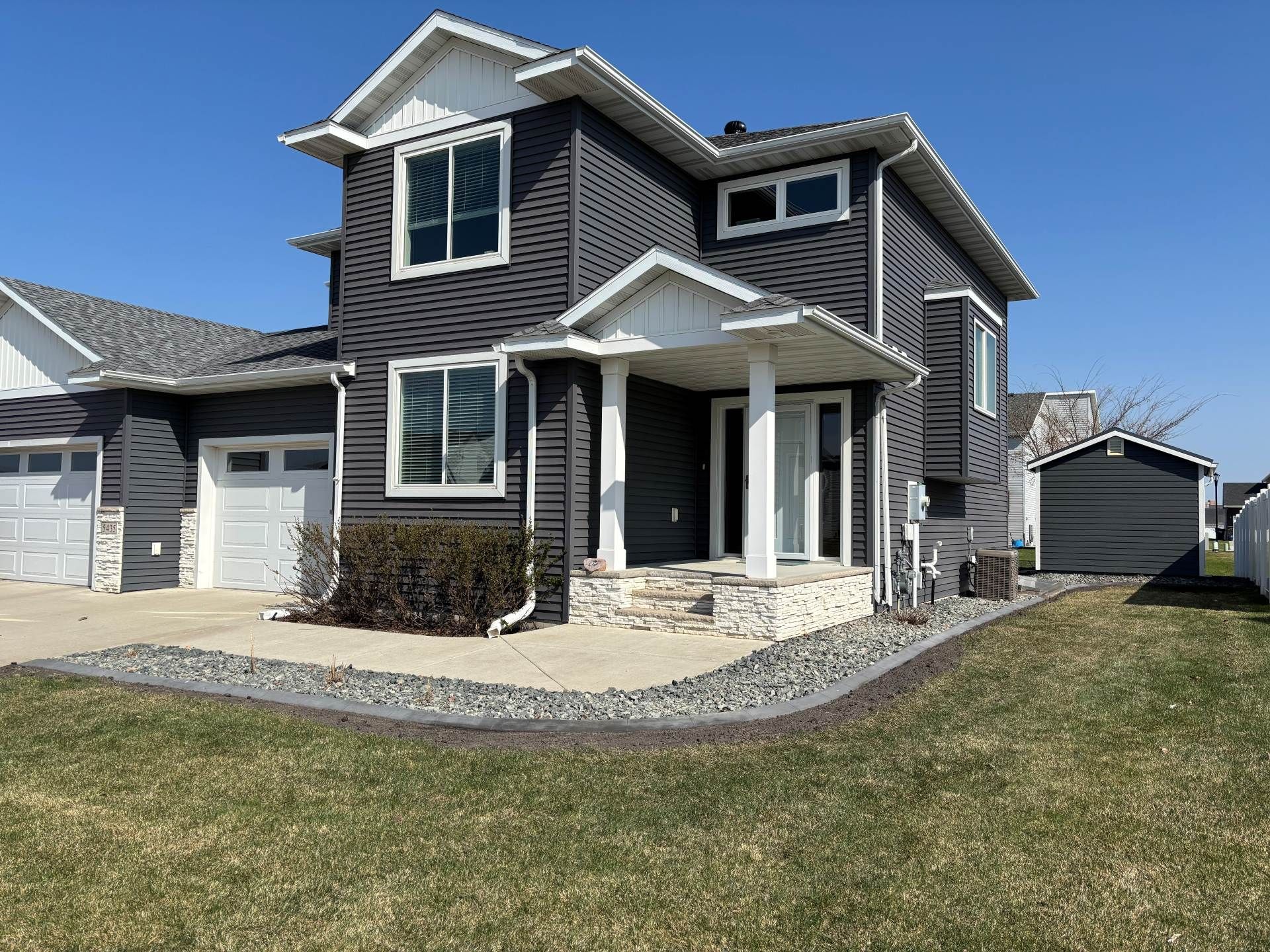 A two-story dark gray house with a stone-accented porch, a white front door, and a detached shed on a sunny day.
