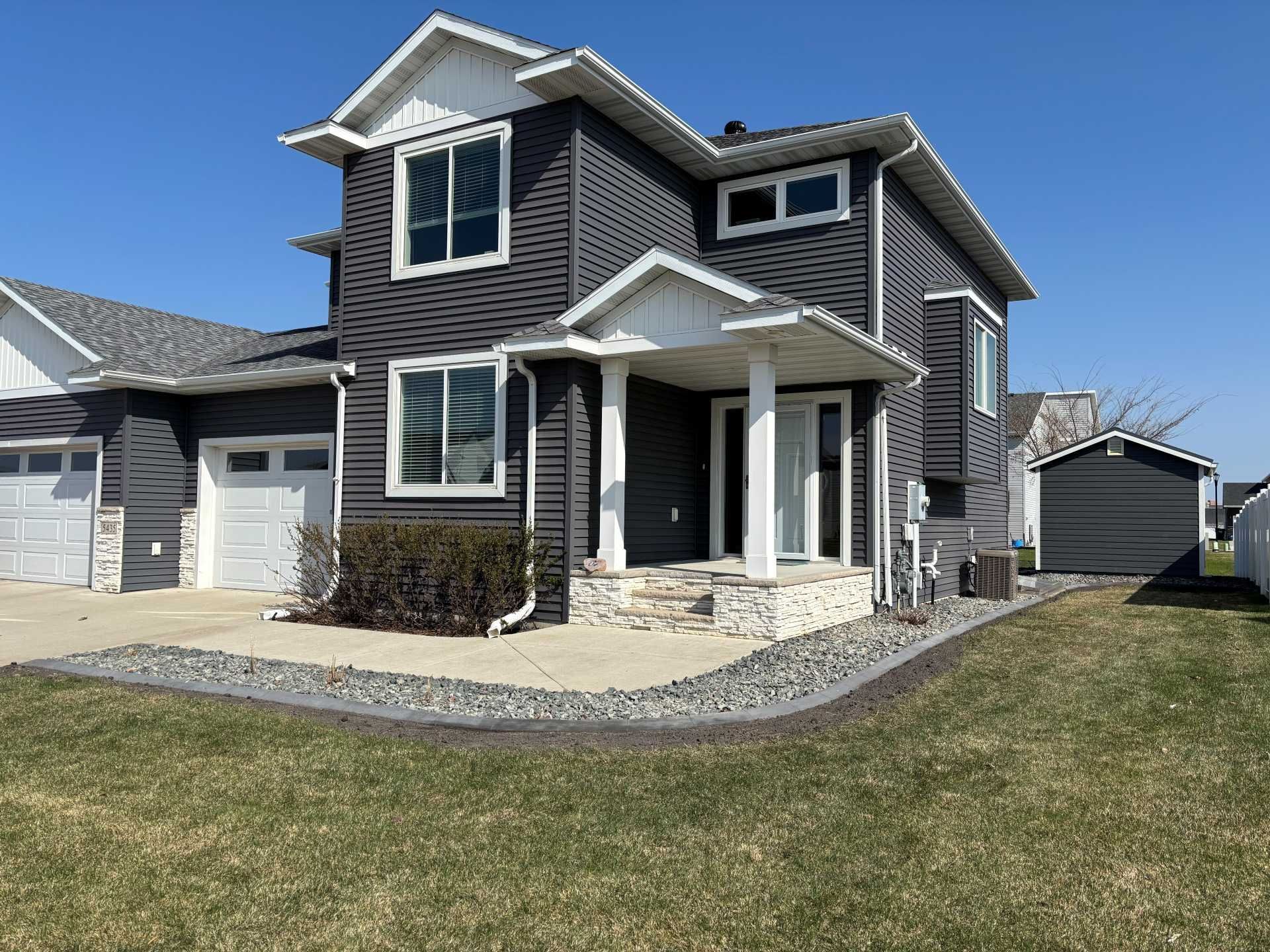 A two-story house with dark gray siding, a stone base, a white porch, and an attached two-car garage under a blue sky.