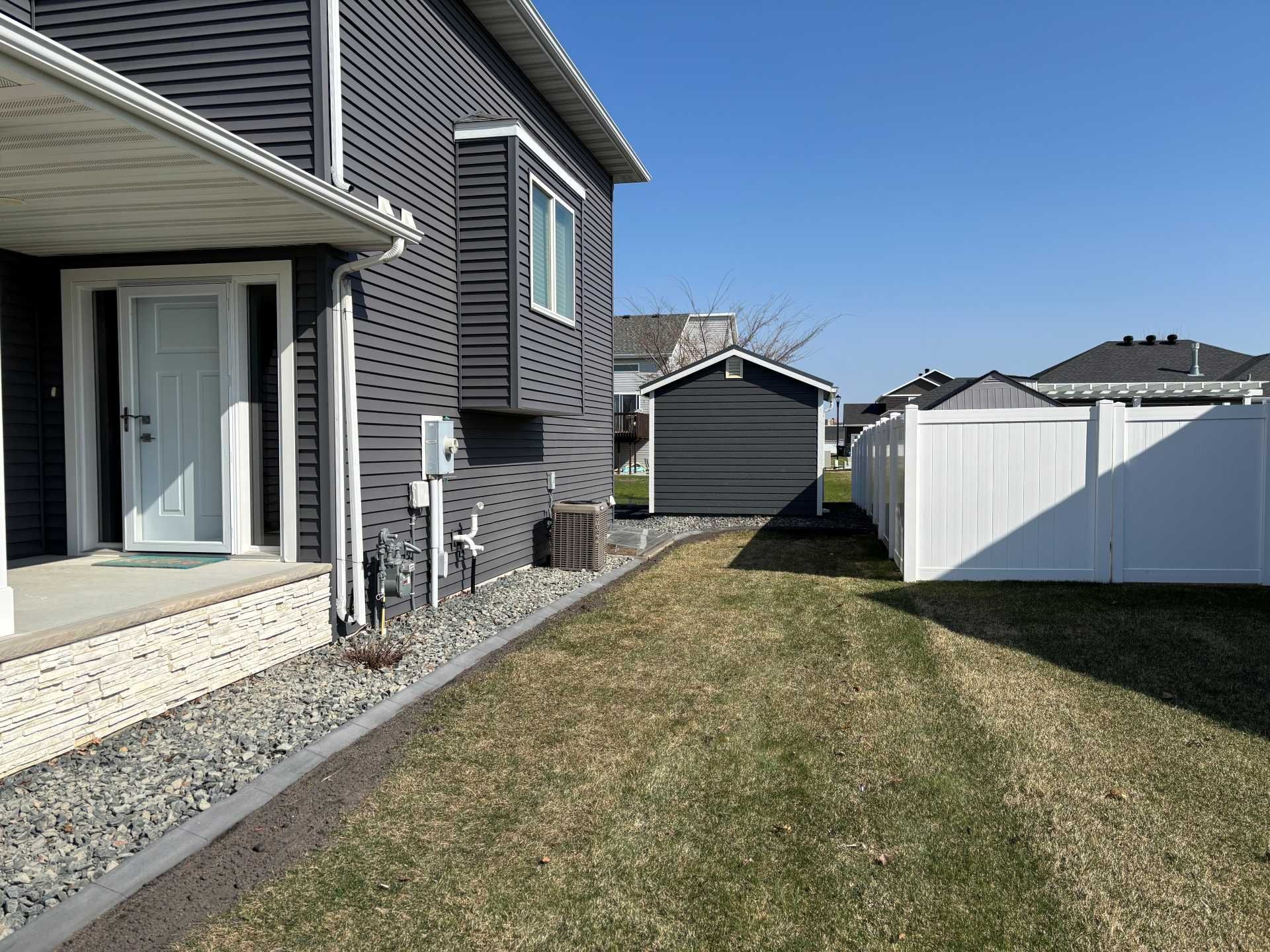 A gray house with white trim, a porch, and a gravel border next to a lawn, a shed, and a white fence under a blue sky.