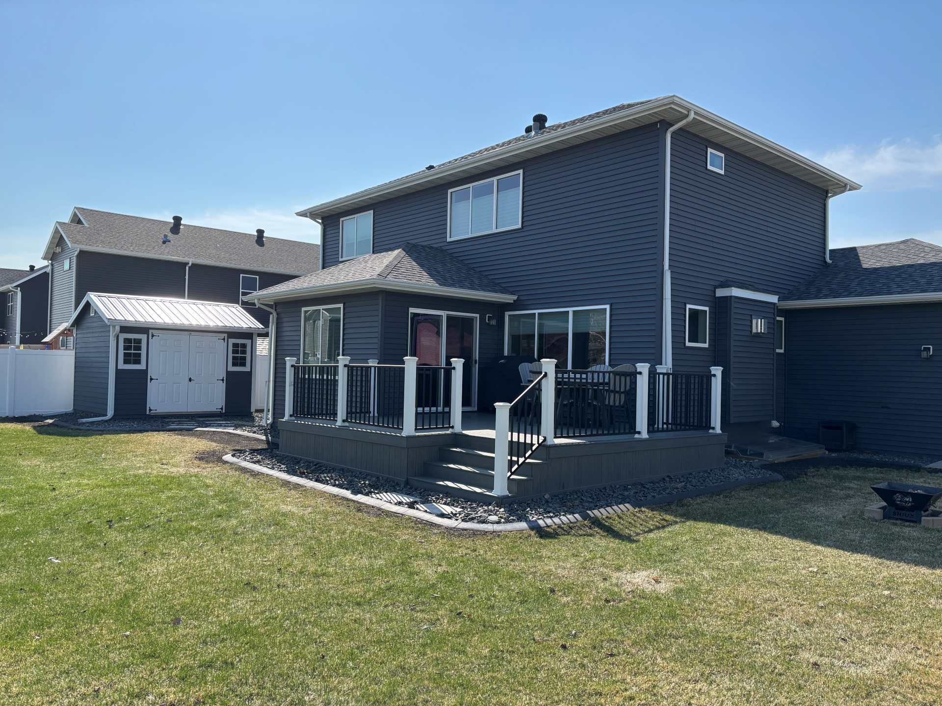 A dark gray two-story house with a wooden deck and a small white shed in the backyard on a sunny day.