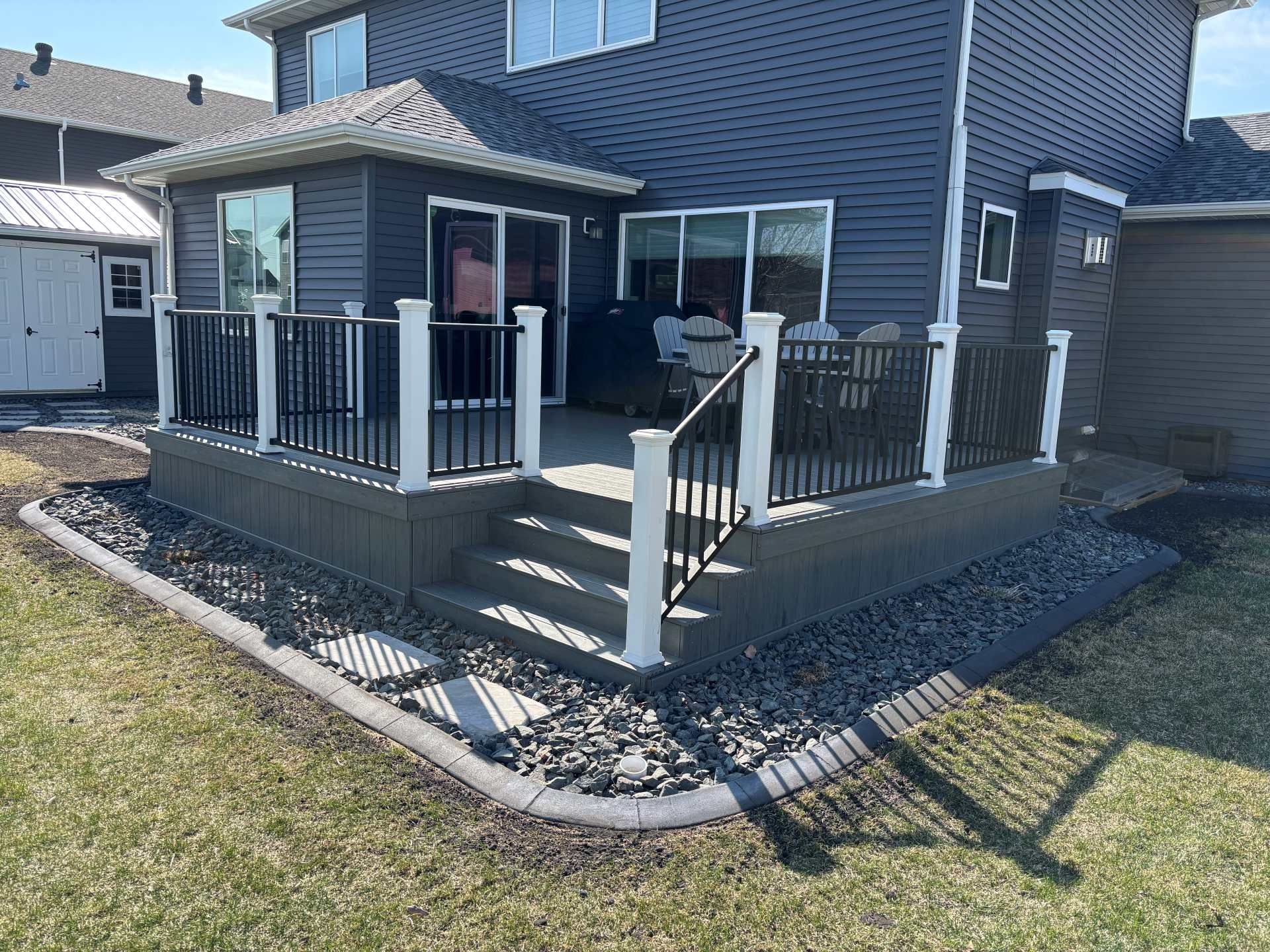 A raised deck with black railings and a white trim border, surrounded by grey decorative stone against a blue house.