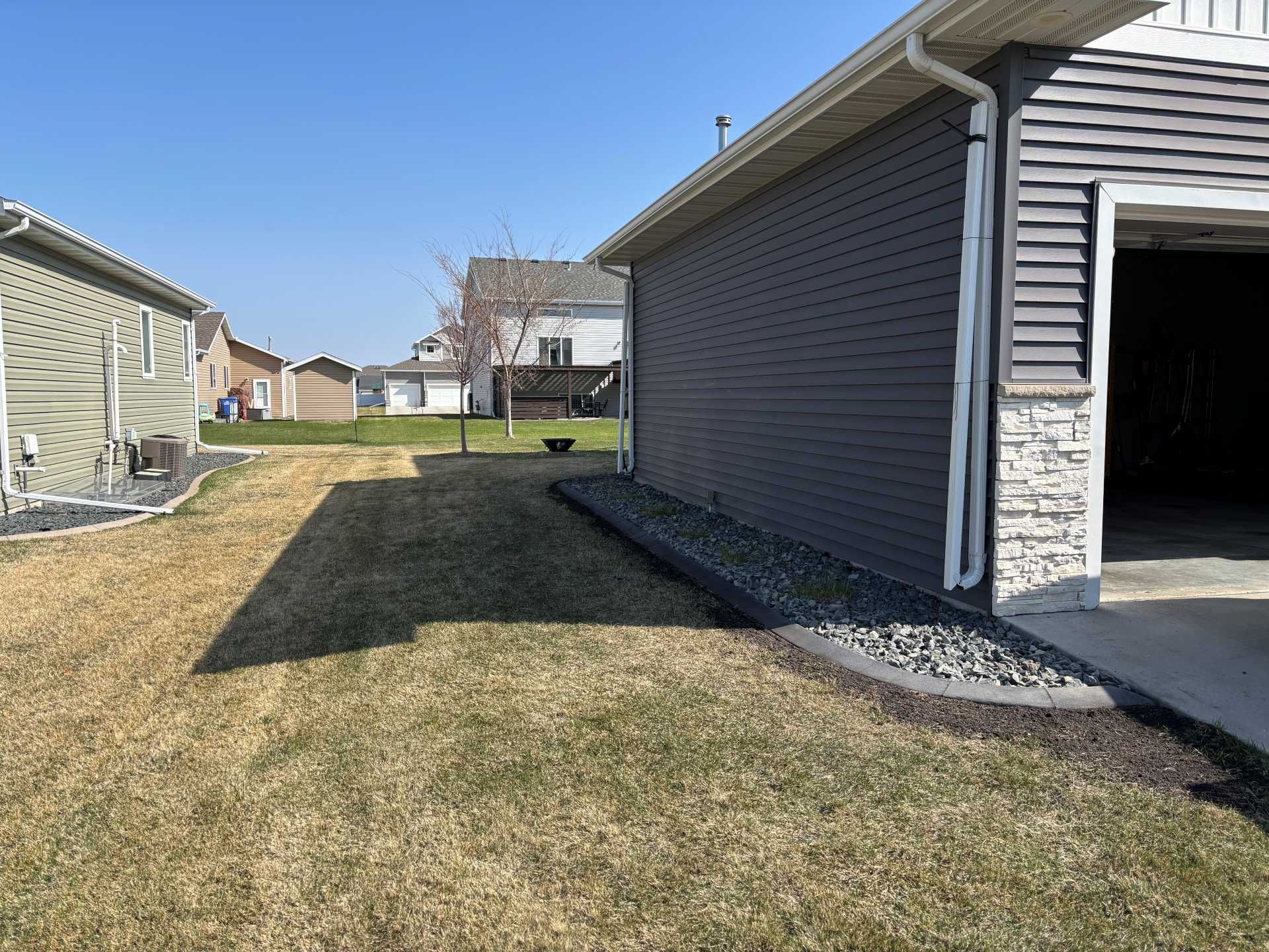 A side view of a house with dark gray siding, a stone base, and a rocky landscape border next to a dry grassy yard.