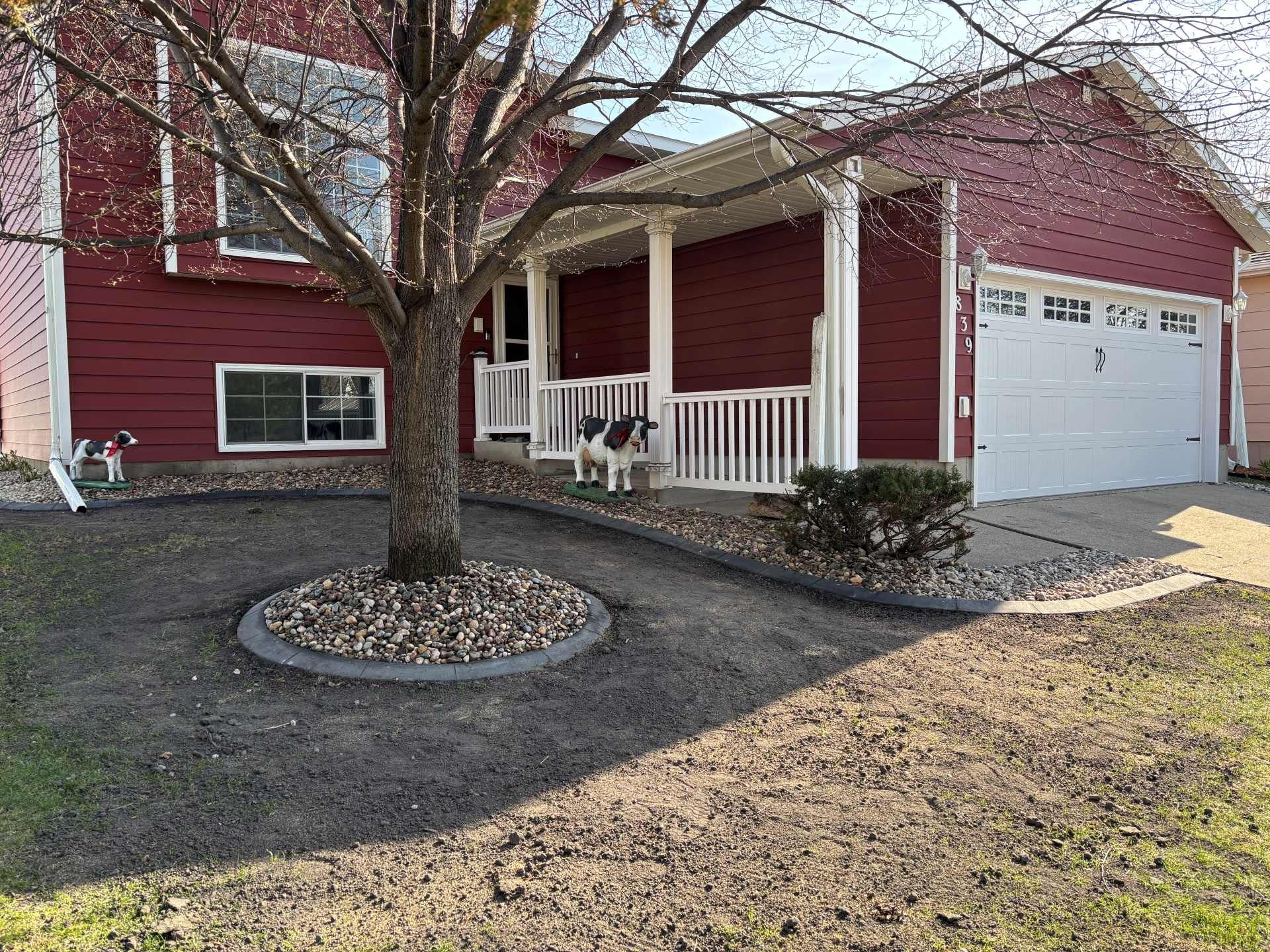 A red house with a white garage and porch, a central tree with a stone ring, and a small dog standing on the lawn.