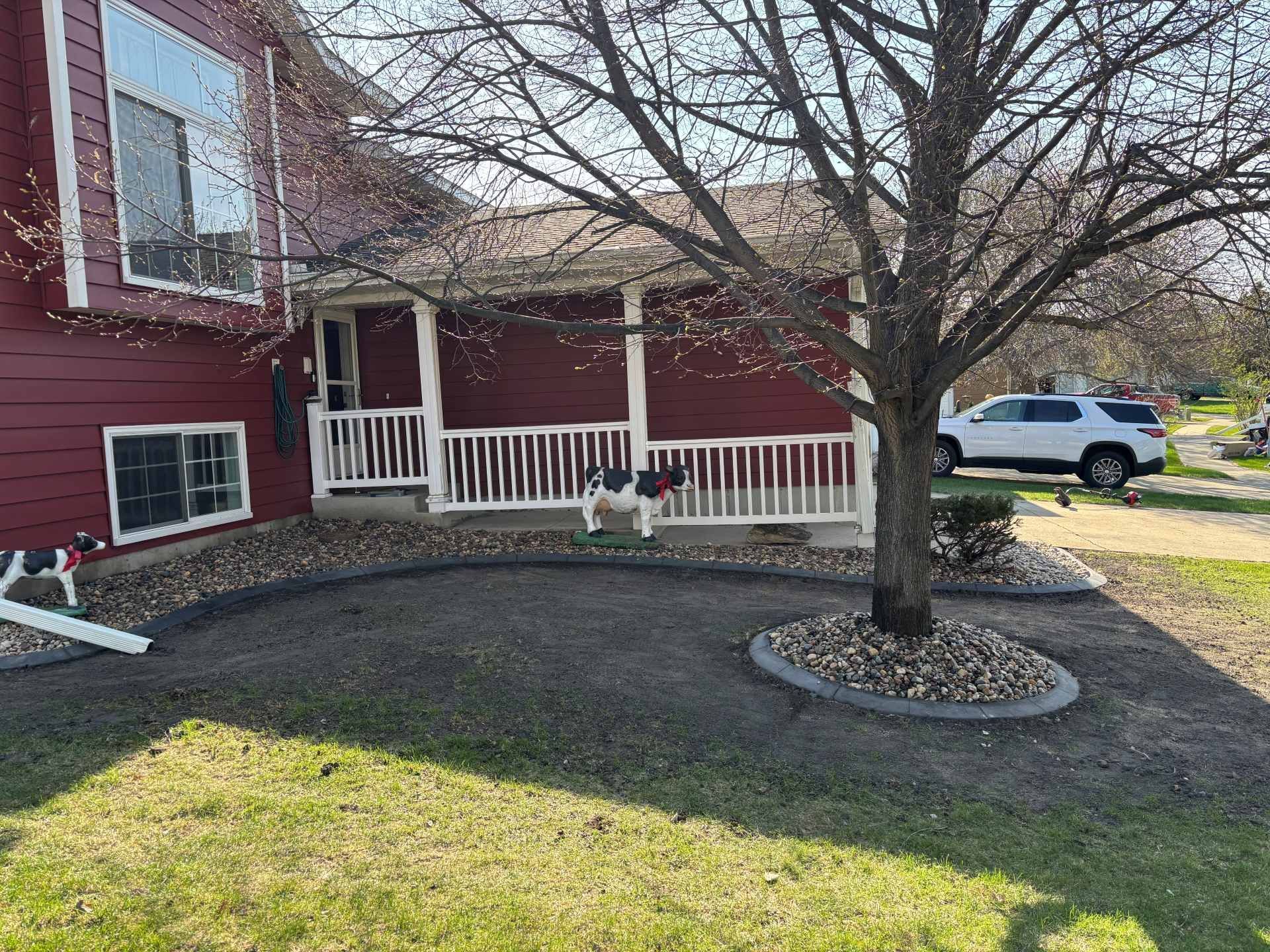 A red house with a porch and a gravel-filled garden bed beneath a tree, with dog lawn ornaments on the grass.