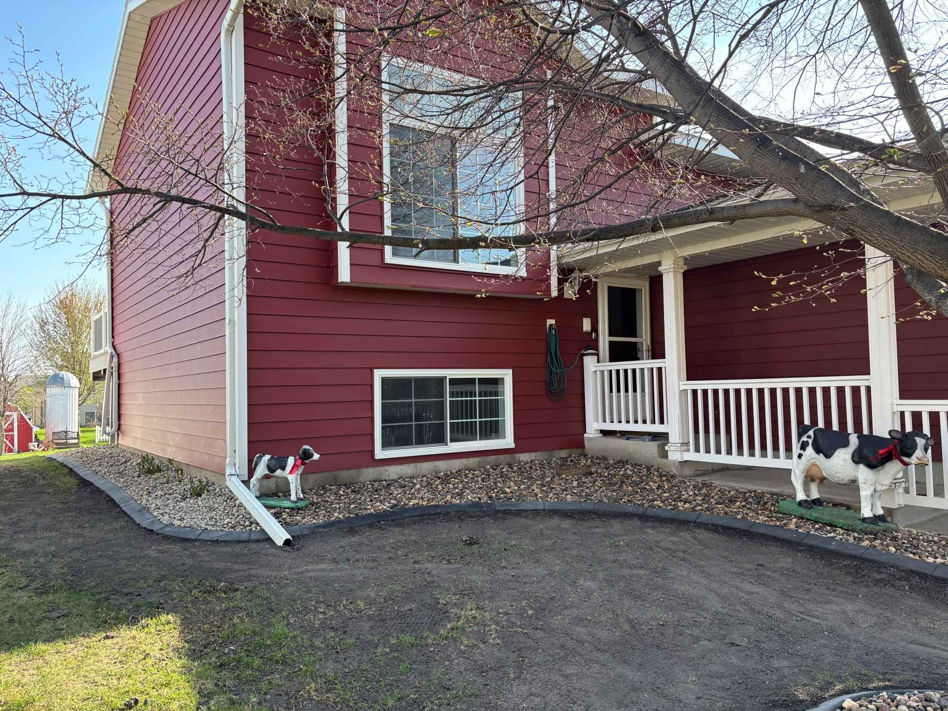 A red house with white trim, a porch, gravel landscaping, and decorative cow and dog statues in the front yard.