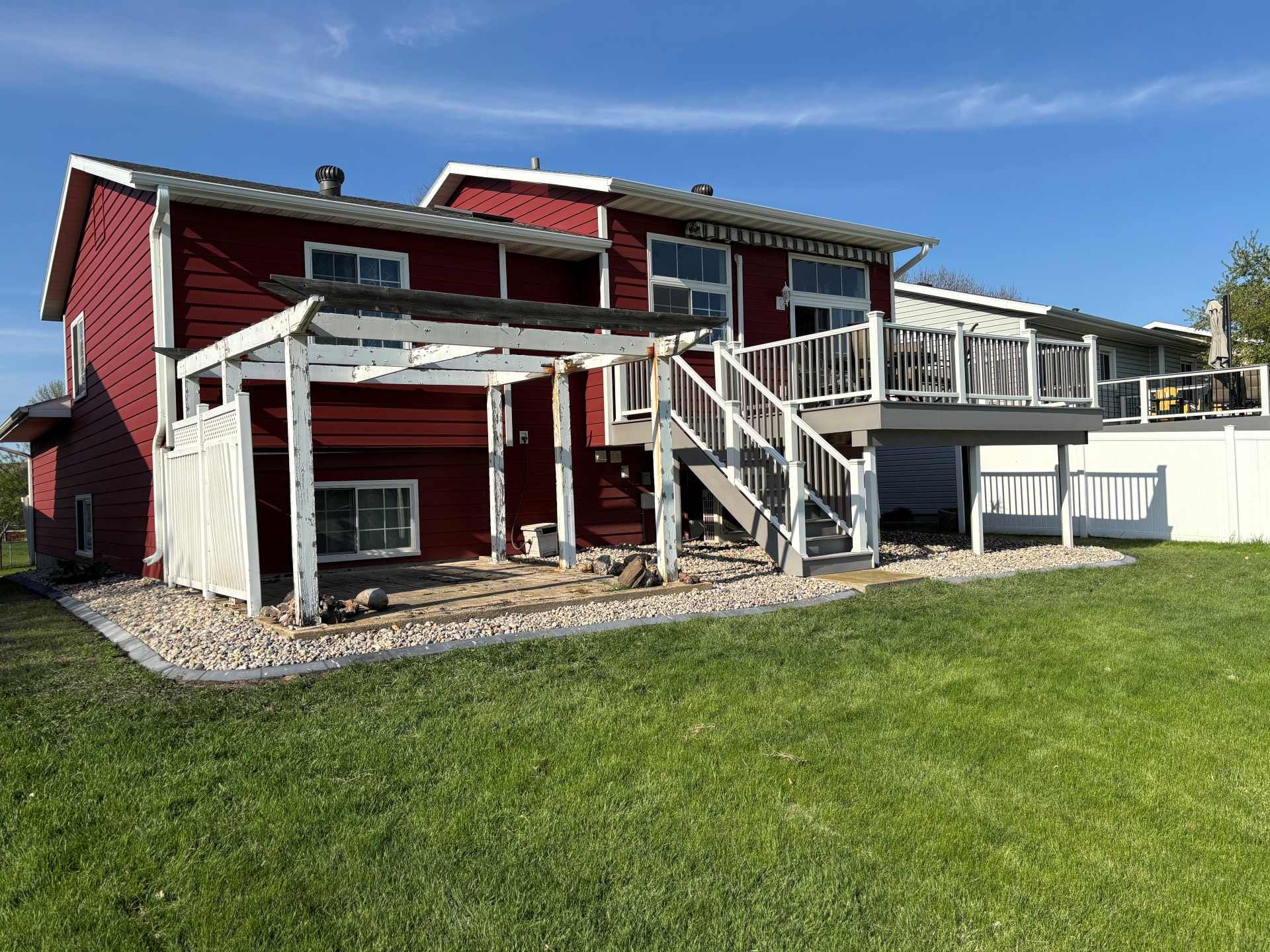 A two-story red house with a white wooden pergola, a raised deck, stairs, and a gravel patio in a grassy backyard.