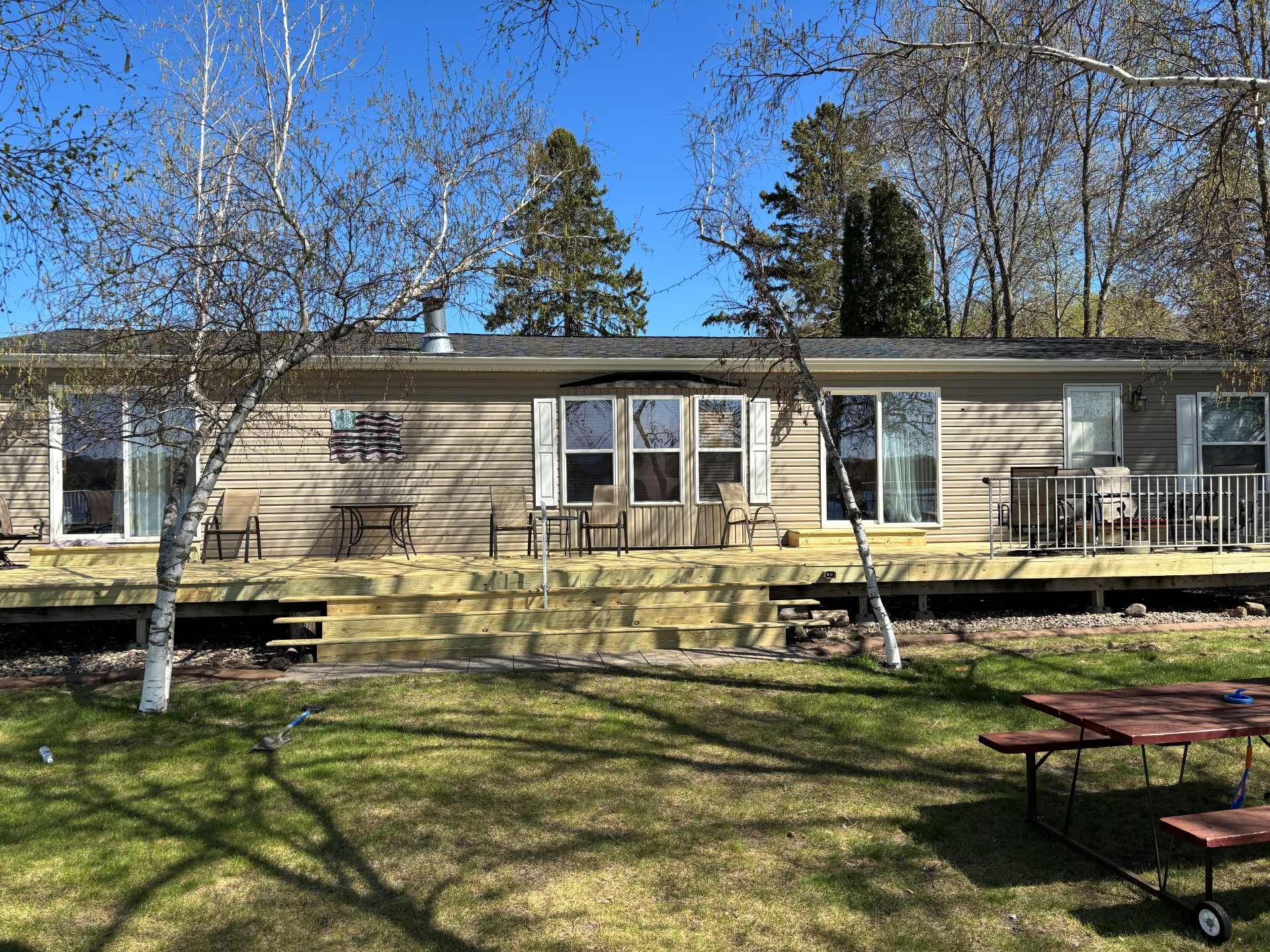 A single-story tan house with a wide wooden deck and glass sliding doors, set behind a lawn with a picnic table.