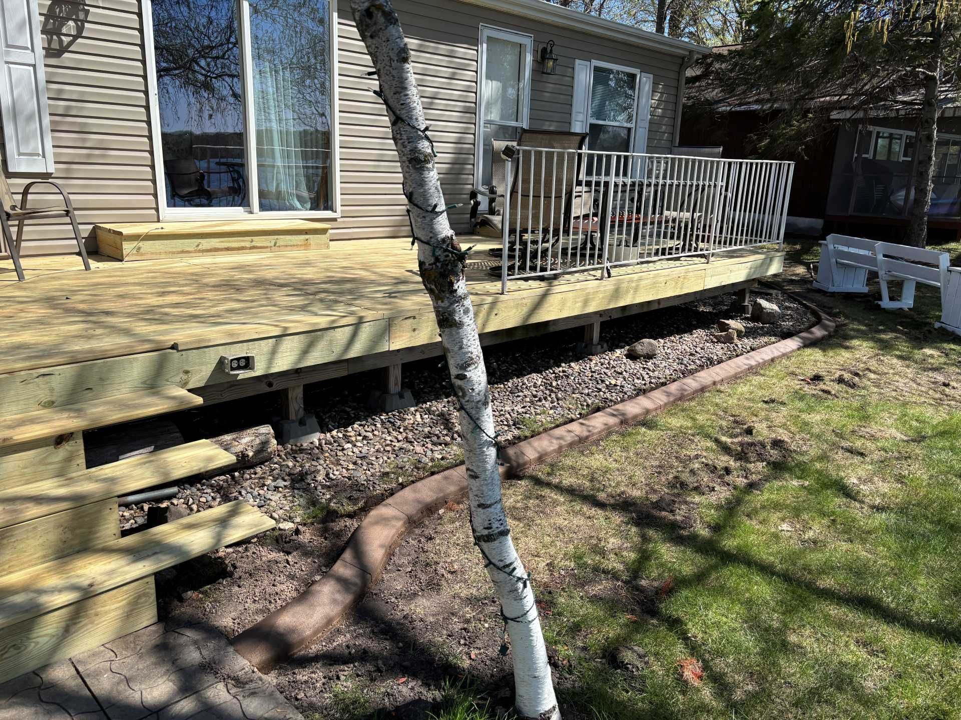 A new wooden deck attached to a house with a white birch tree in the foreground and a gravel bed along the base.