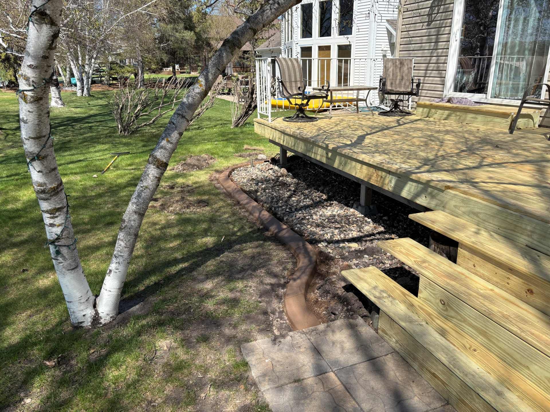 A wooden deck staircase sits next to a landscaped garden bed with a curved concrete border and scattered leaves.