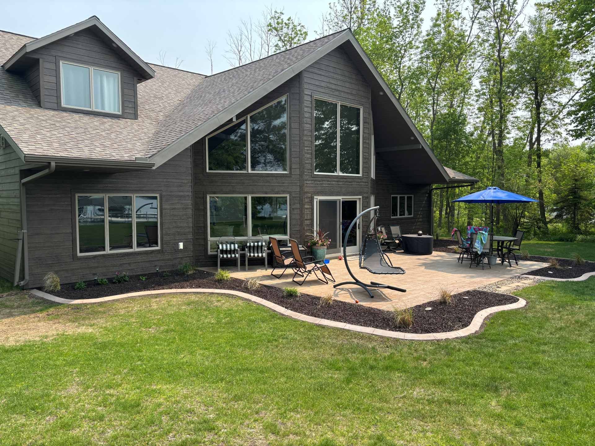 A dark brown house with a large patio, outdoor furniture, a blue umbrella, and a curved landscaped garden bed.