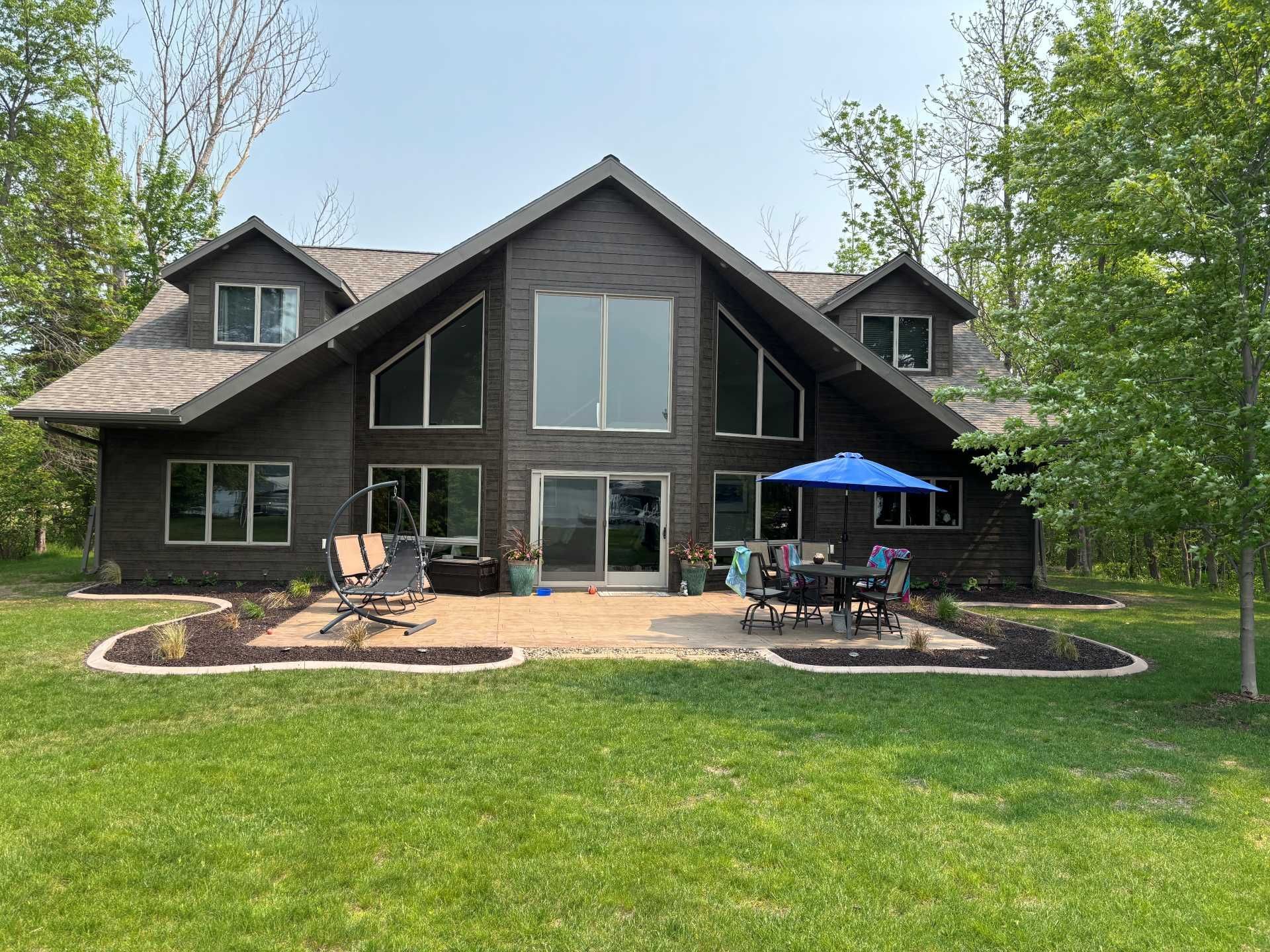 Dark wood, A-frame house with large windows, a stone patio, patio furniture, and a blue umbrella, set in a grassy yard.