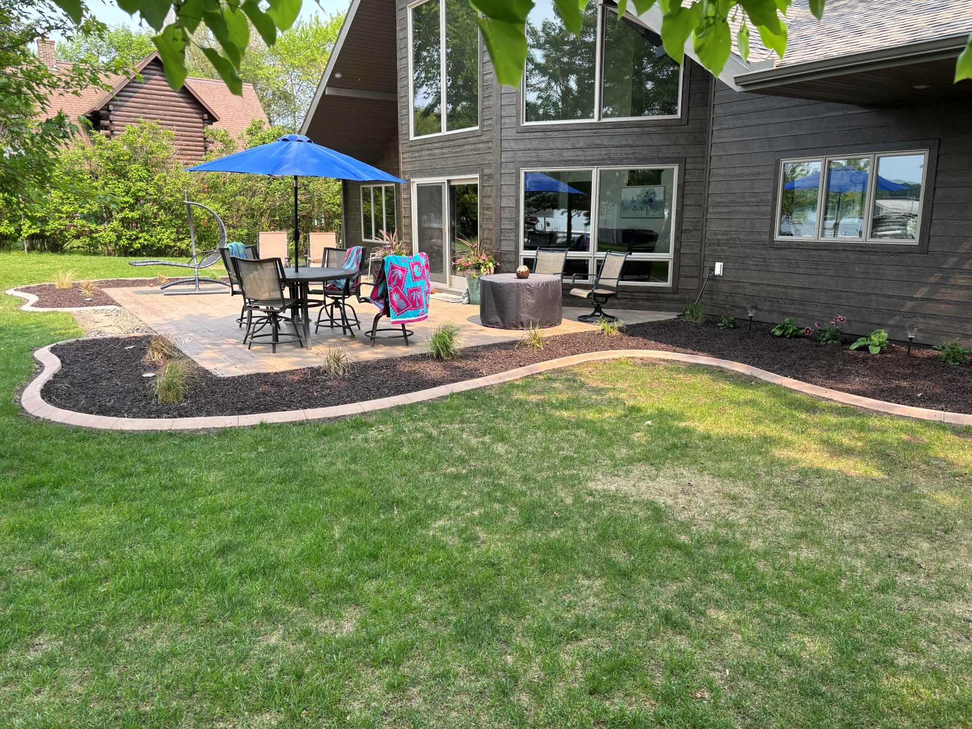 A back patio with a blue umbrella, dining set, and fire pit sits next to a dark wood house overlooking a grassy lawn.