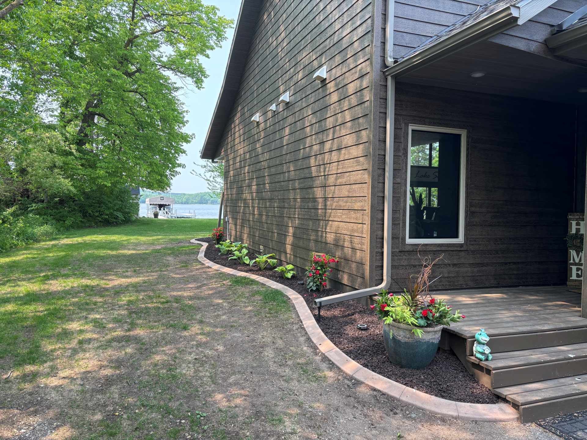 Dark wood siding building with a curved garden bed of hostas and a potted plant next to wooden steps overlooking a lake.